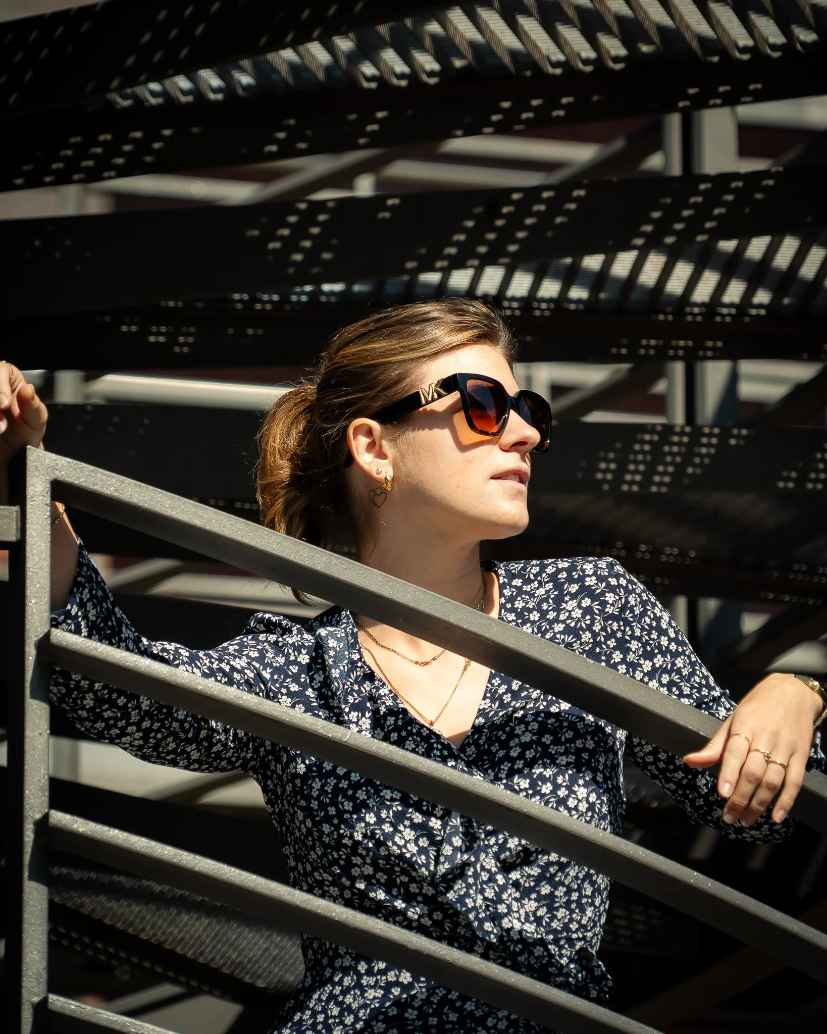 Eine Frau mit Sonnenbrille und blumigem Kleid steht an einem Geländer einer Metalltreppe, beleuchtet von Sonnenlicht, mit Schattenspiel im Hintergrund.