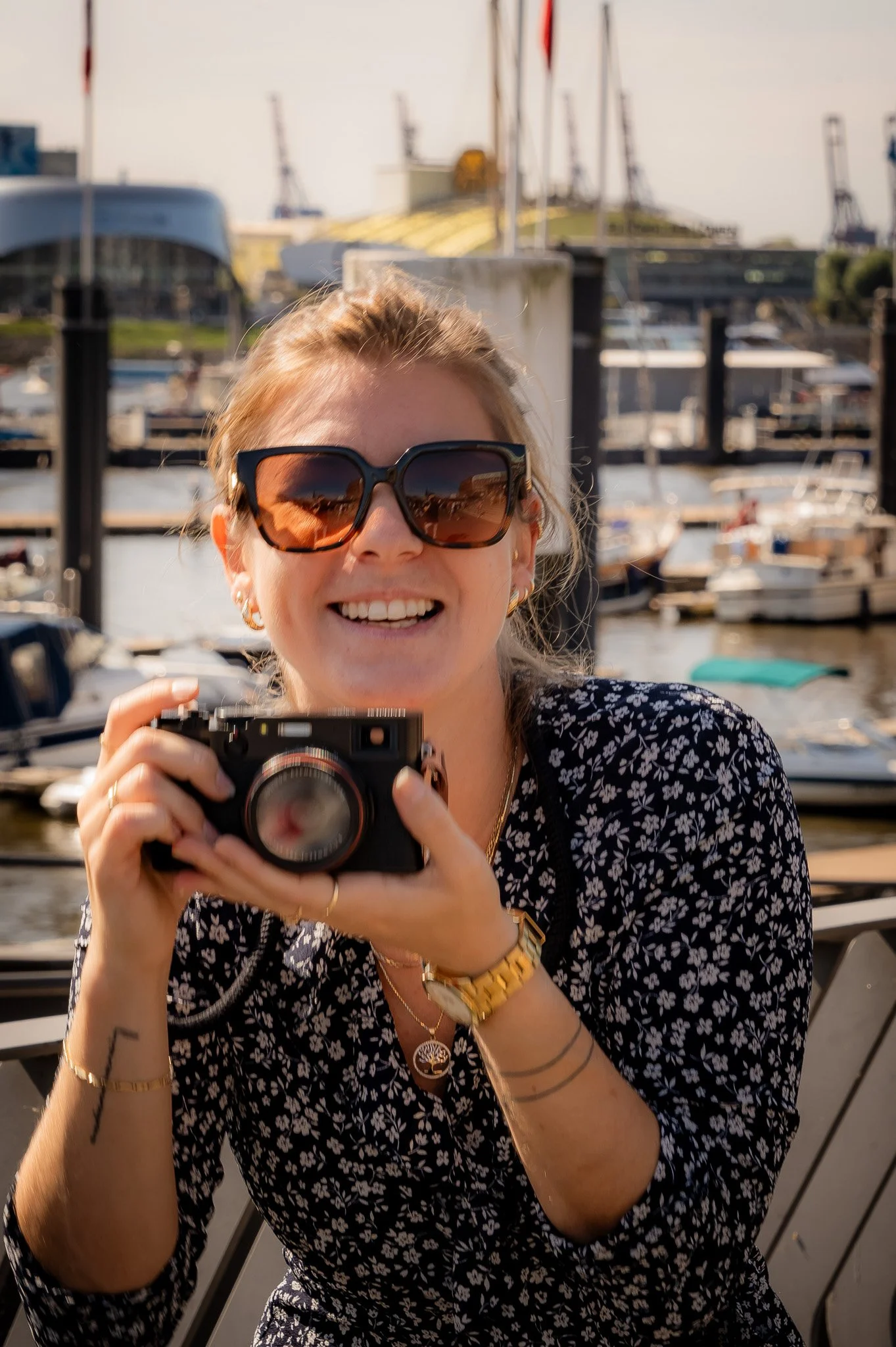 Frau mit Sonnenbrille hält eine Kamera und lächelt, im Hintergrund ein Hafen mit Booten.