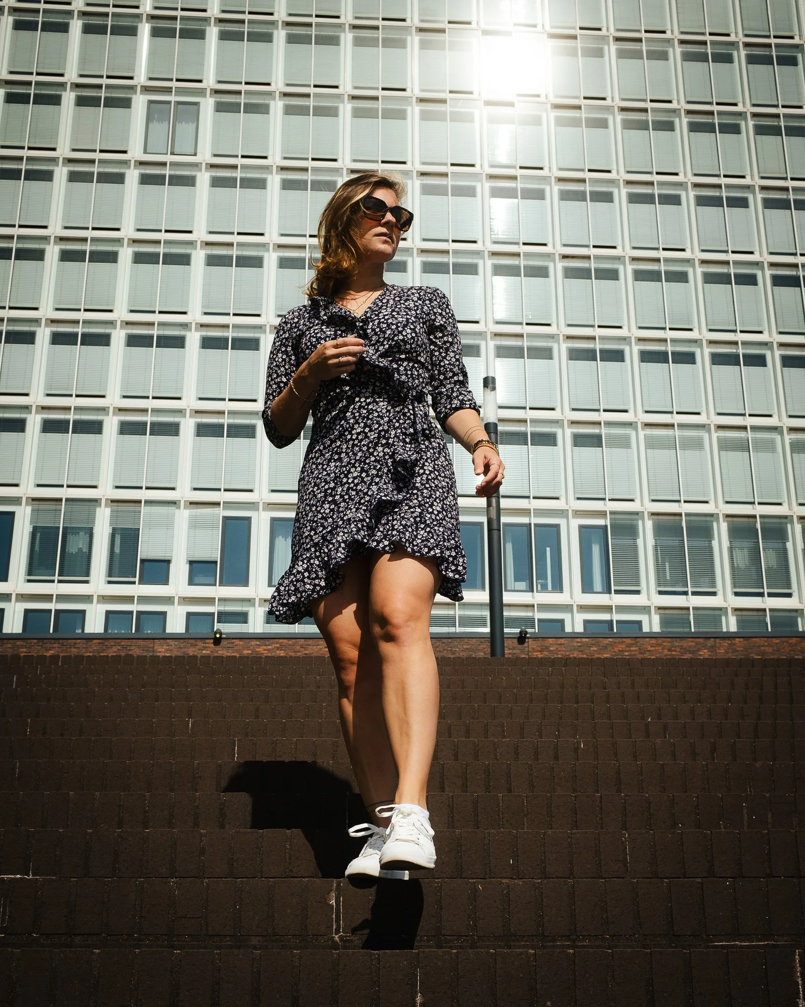Frau in Sommerkleid und Sonnenbrille geht Treppen hinab vor einem Hochhaus.