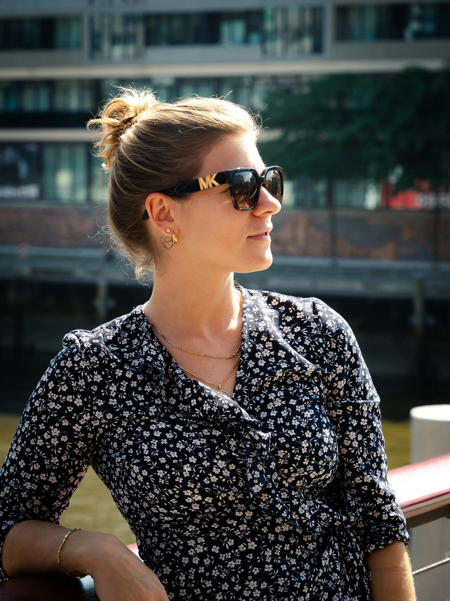 Eine Frau mit Sonnenbrille und hochgestecktem Haar, die ein dunkelblaues Kleid mit weißen Blumen trägt, steht an einem Geländer. Im Hintergrund ist ein Gebäude zu sehen.