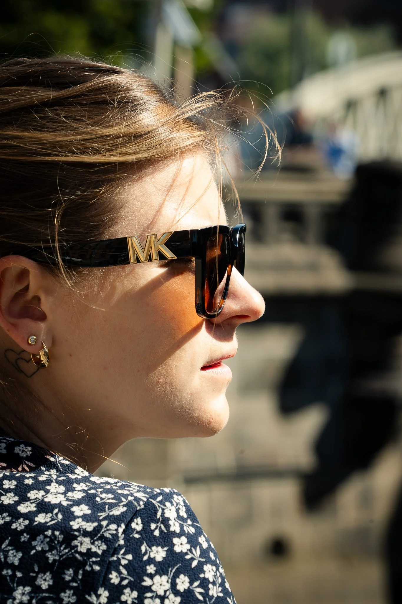 Nahaufnahme einer Frau mit Sonnenbrille und Ohrringen, Sonnenlicht im Hintergrund. Seitliche Ansicht.
