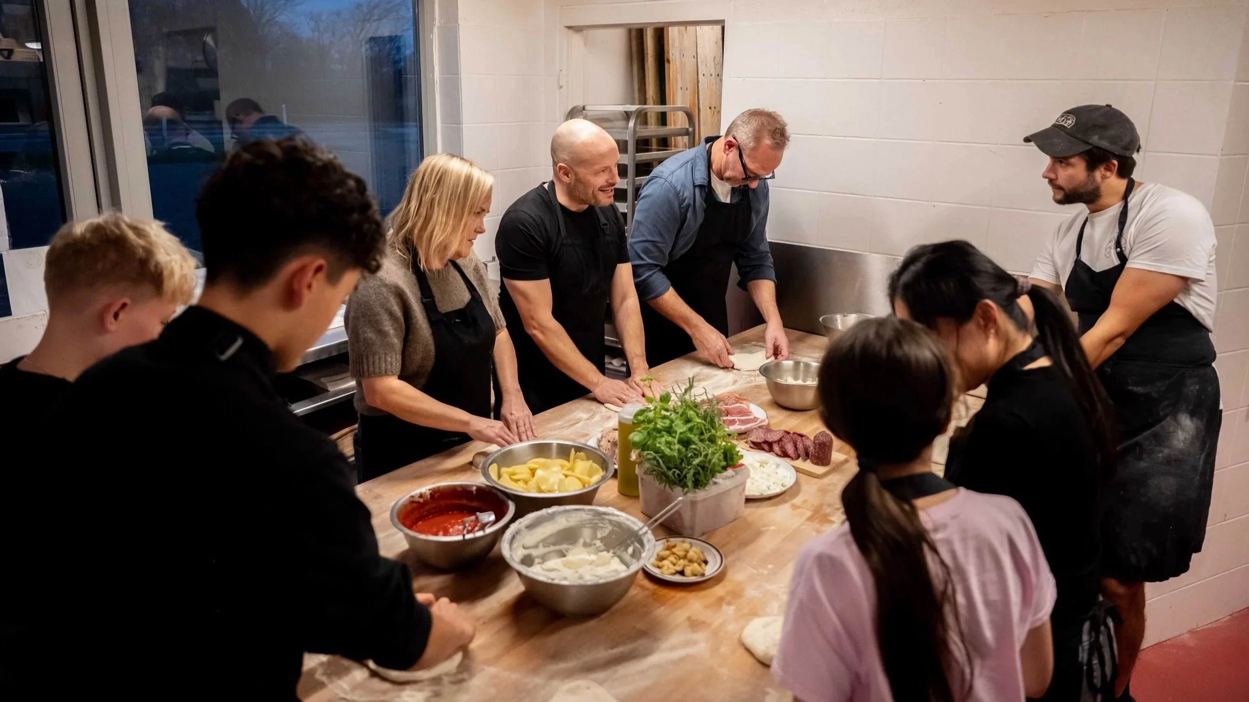En gruppe mennesker, inklusive voksne og børn, er samlet omkring et træbord, hvor de laver mad sammen. Der er forskellige ingredienser som spaghetti, kød, og grøntsager på bordet.