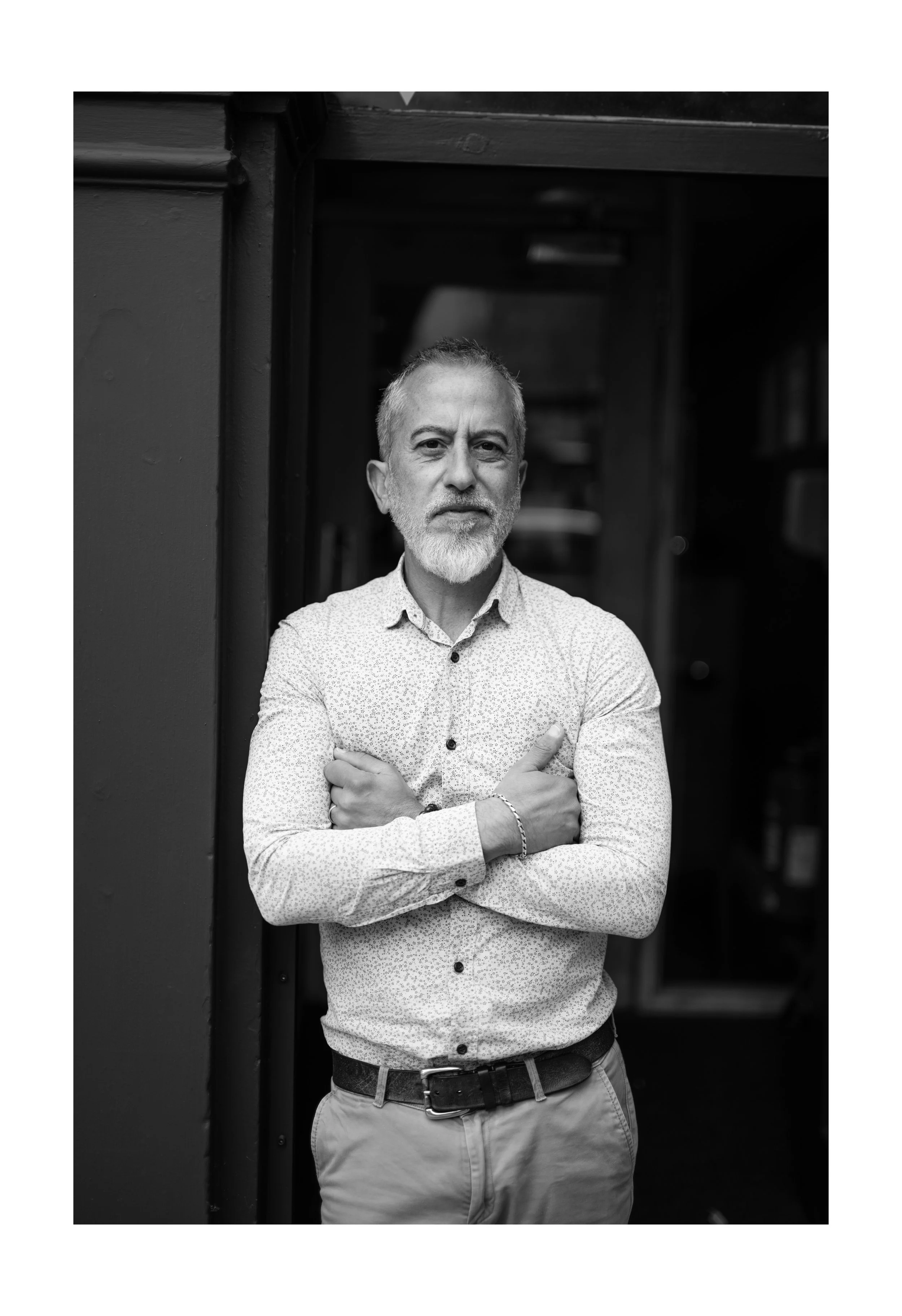 Black and white photo of a confident middle-aged man with short gray hair, beard, and mustache, wearing a patterned button-up shirt with sleeves rolled up and crossed arms, standing outside near a structure with dark walls.