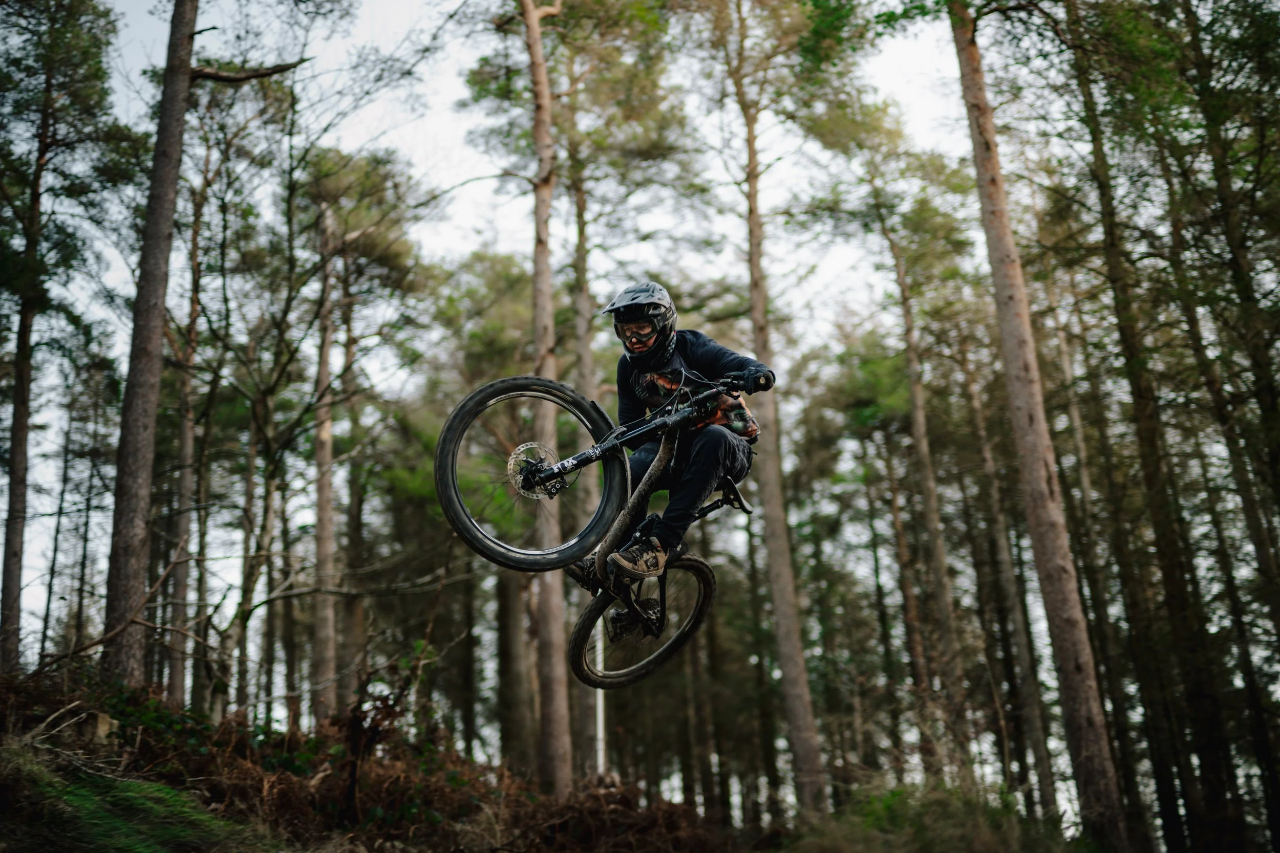 A mountain biker wearing a helmet and protective gear rides off a dirt trail in a forest with tall trees and dense foliage.