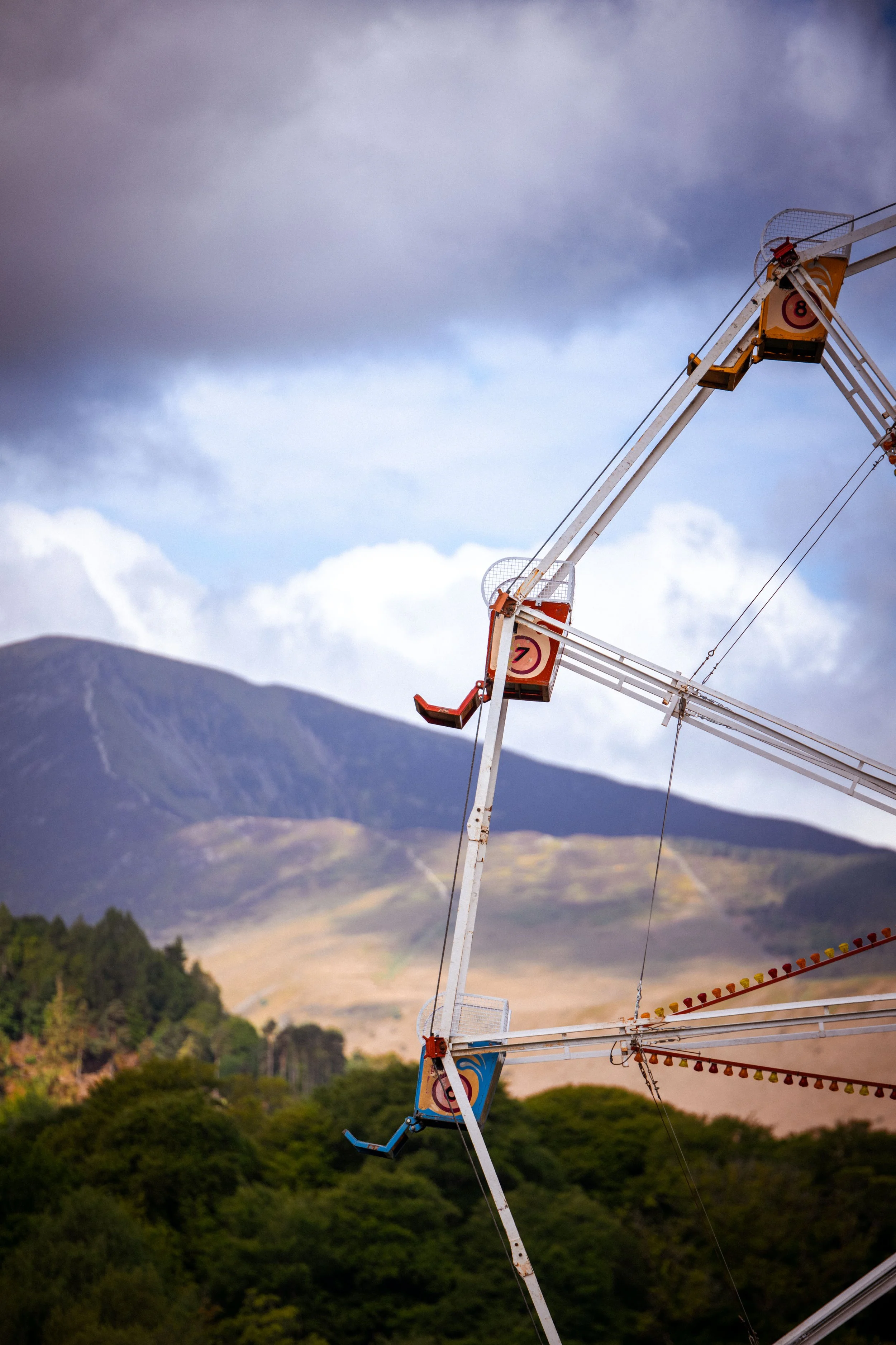 A derelict amusement park ride with a ferris wheel, showing signs of rust and disrepair, against a backdrop of cloudy sky and distant mountains.