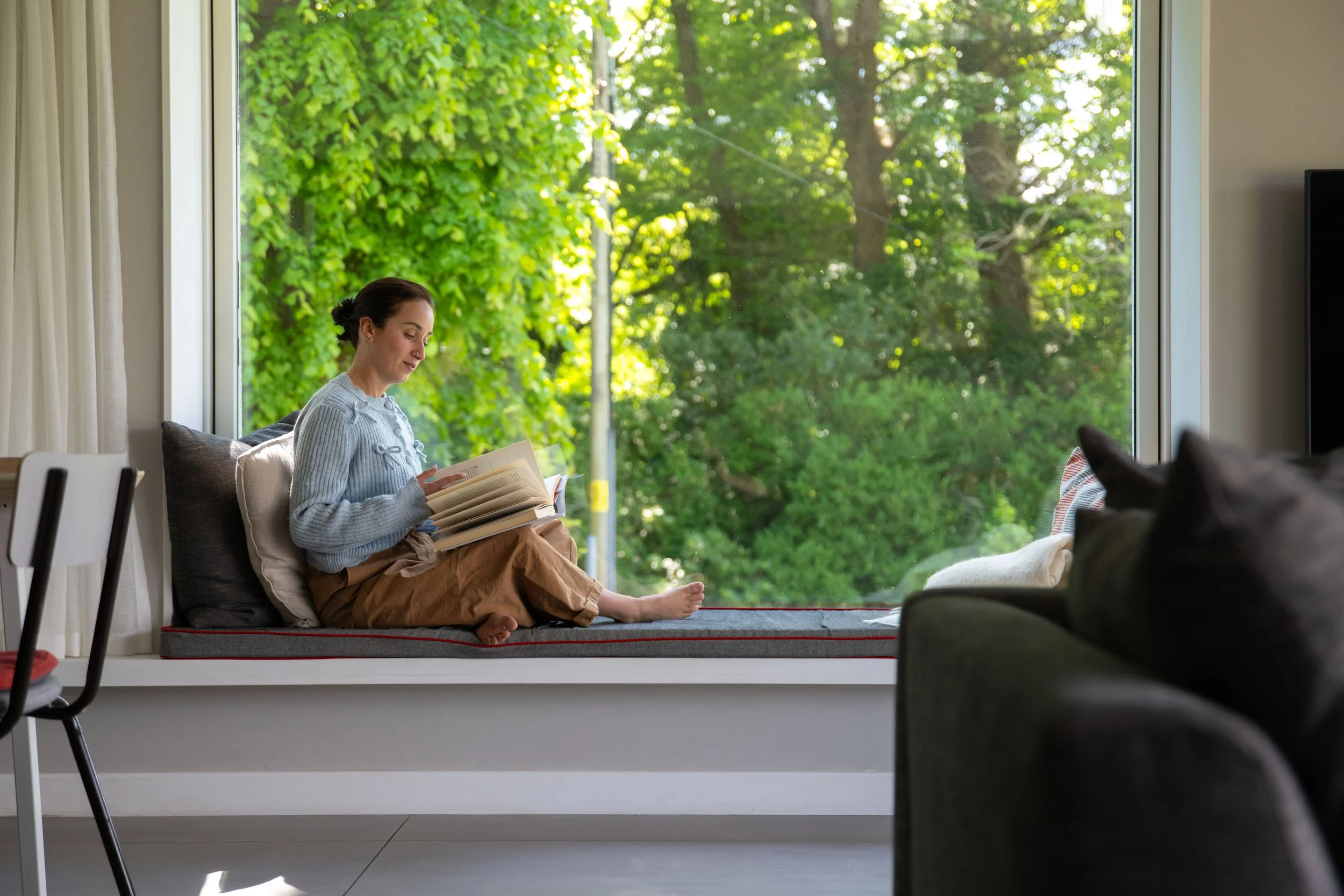 A woman sitting on a window seat reading a book, with greenery outside visible through a large window.
