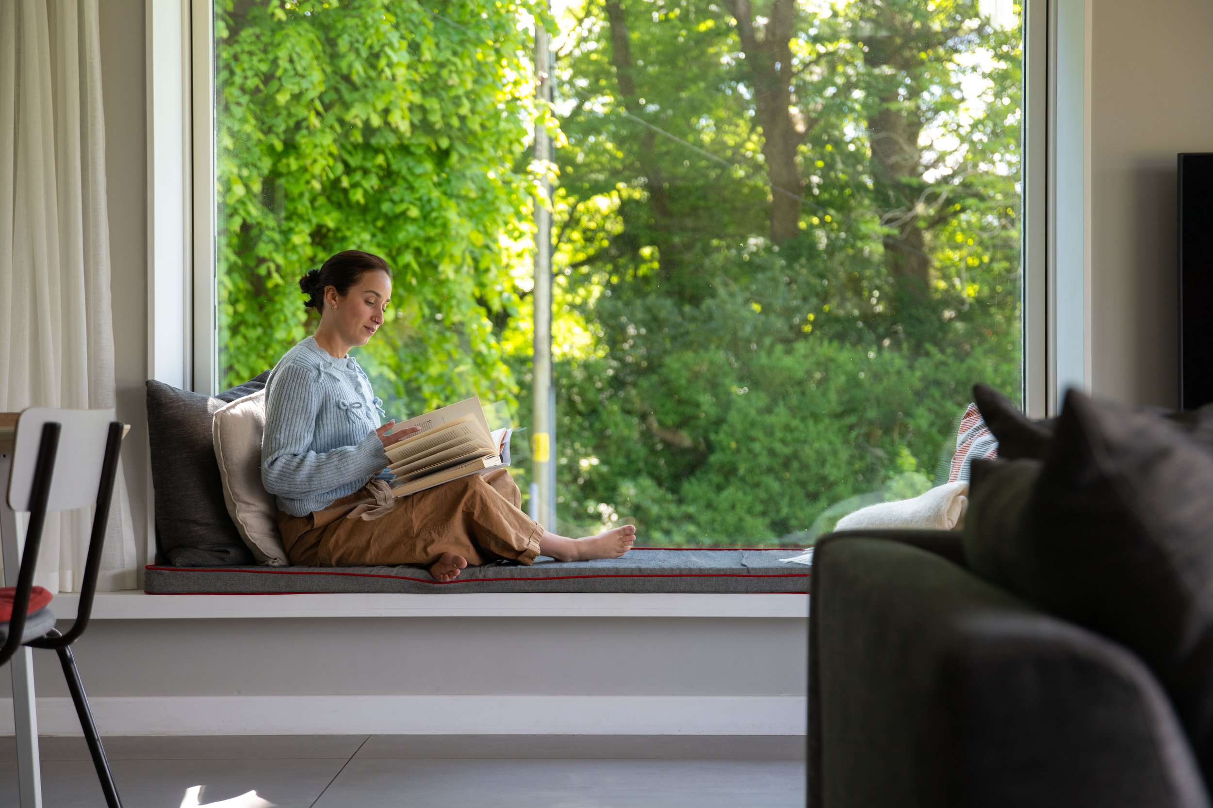 A woman sitting on a window seat reading a book, with greenery outside visible through a large window.