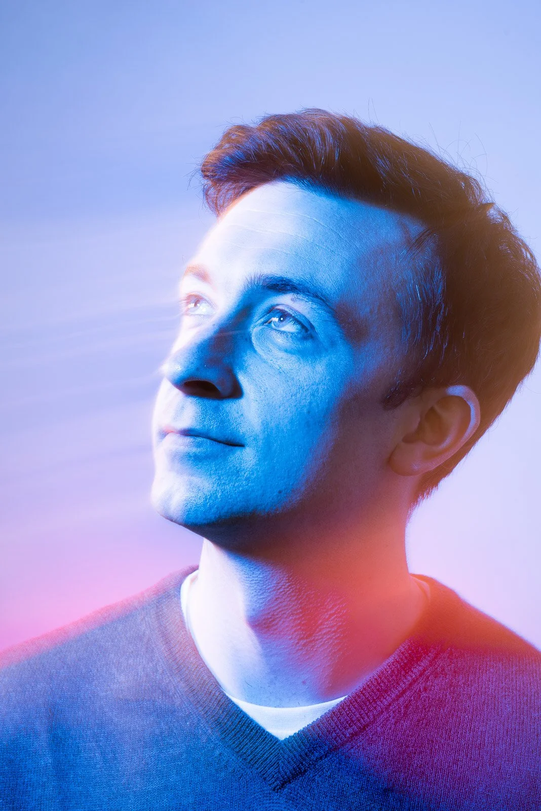 A young man with short dark hair gazing upward with a serene expression, illuminated by blue and pink studio lighting.