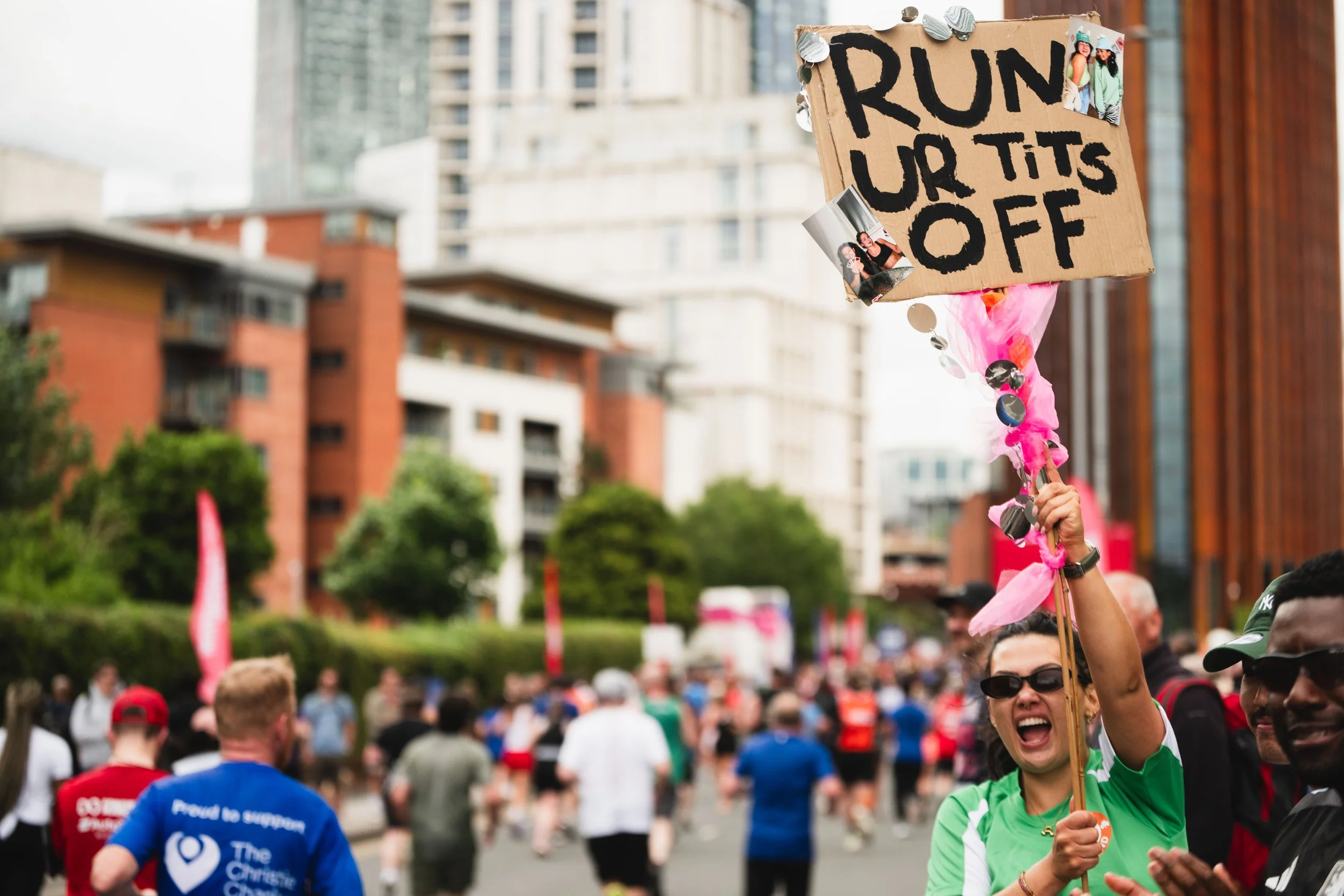 A woman holding a cardboard sign that says 'RUN UR TITS OFF' during a marathon or running event, with a crowd of runners and city buildings in the background.