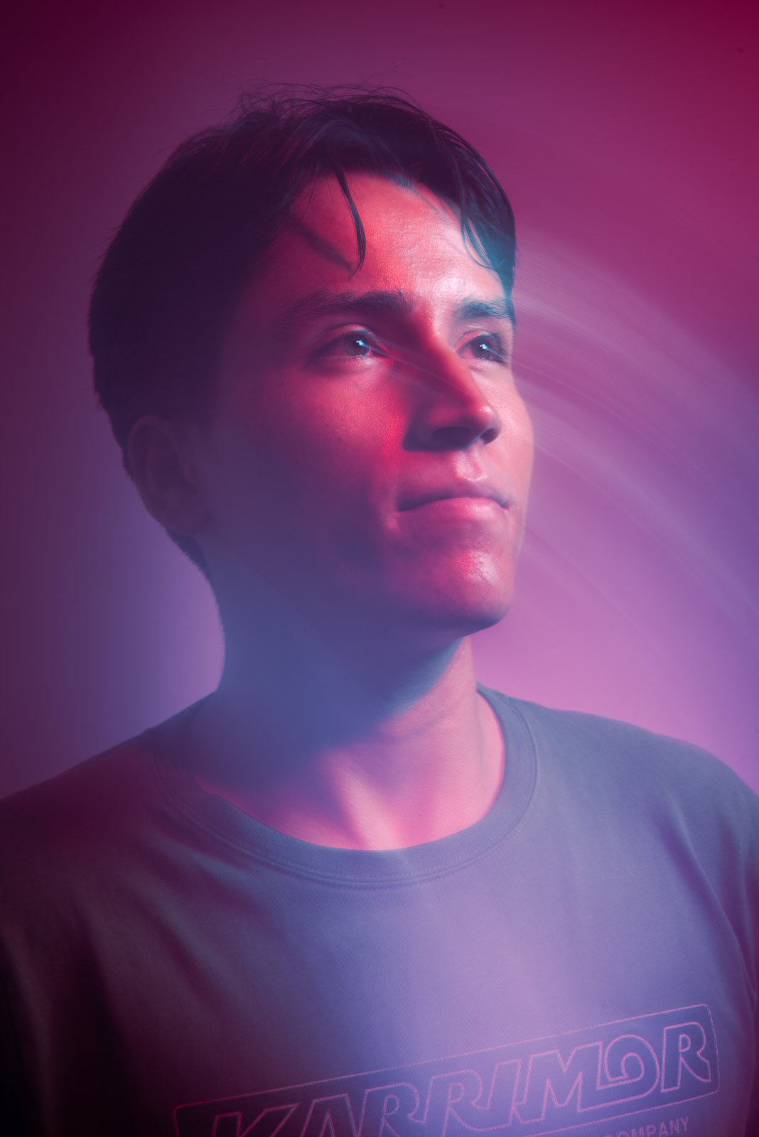 A young man with dark hair looks upward against a pink and purple lit background.