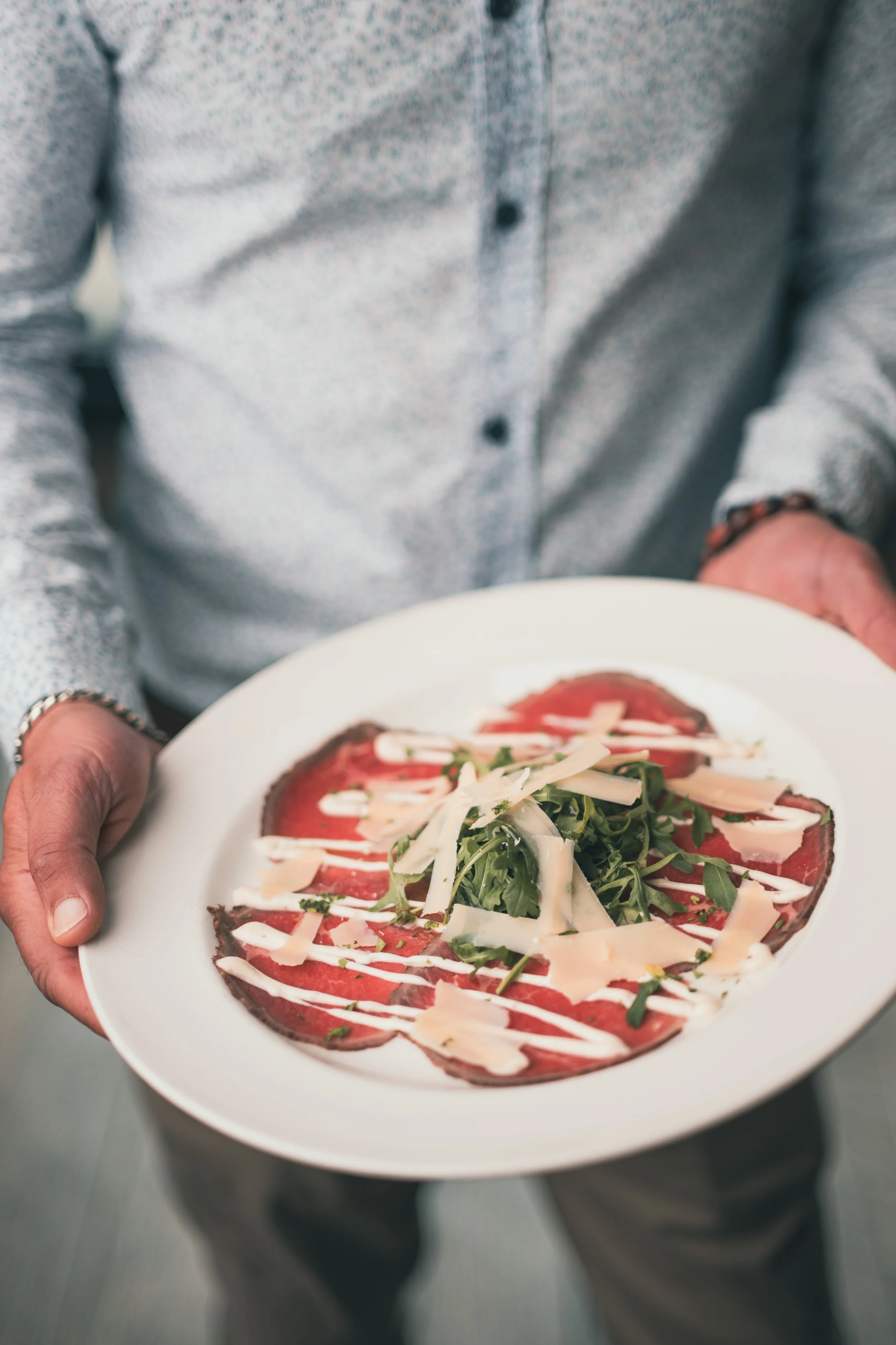 Person holding a white plate with a dish that includes sliced beef, arugula, shredded cheese, and drizzled white sauce.