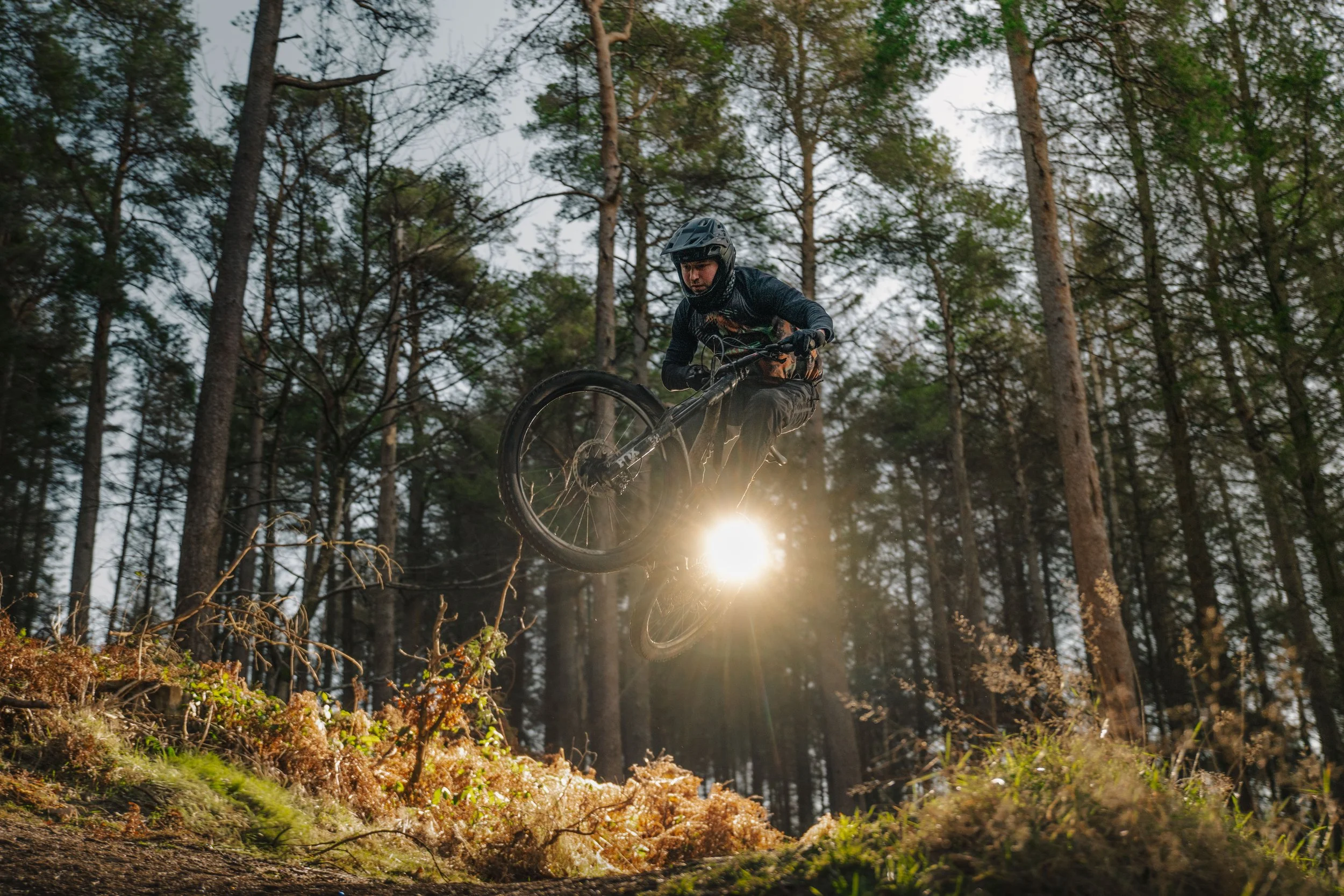 A mountain biker in dark clothing and a helmet jumping over a dirt mound in a forest with tall trees, with sunlight shining through the trees.