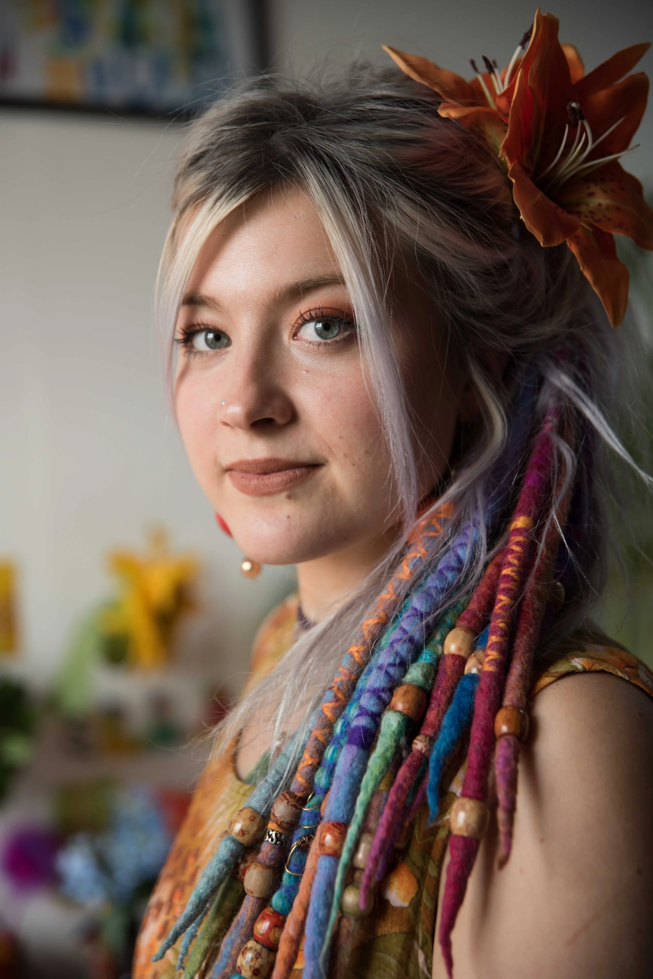 A woman with wavy blonde hair adorned with a large orange lily flower, wearing colorful dreadlocks with beads, and dressed in a patterned scarf.