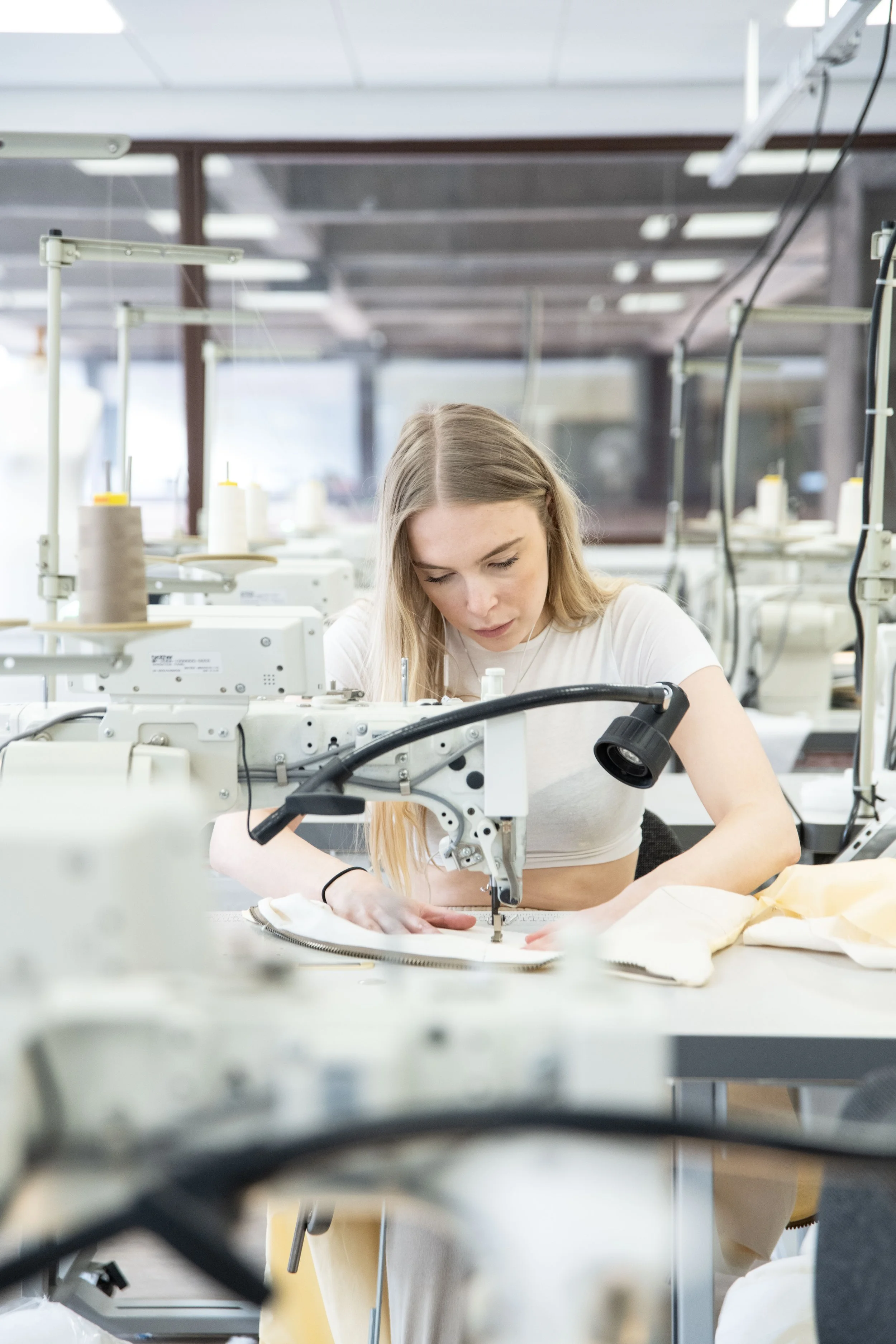 Young woman working with fabric at industrial sewing machine in sewing workshop, with multiple sewing machines and large windows in the background.