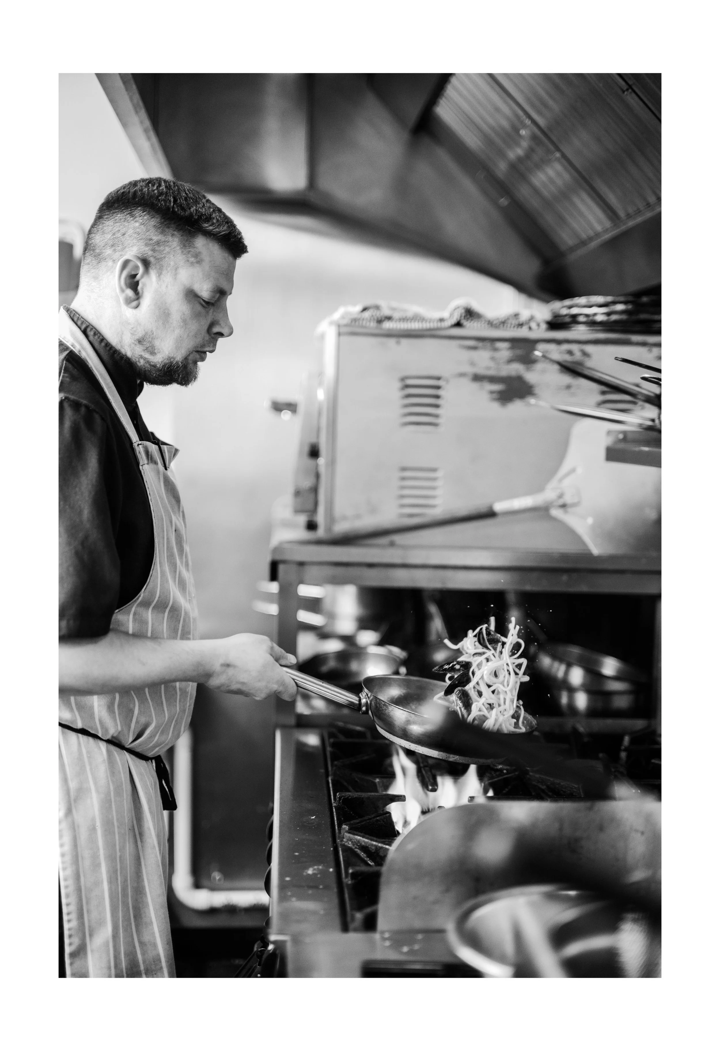 Black and white photo of a man wearing an apron cooking pasta in a commercial kitchen, with stove and pots visible.