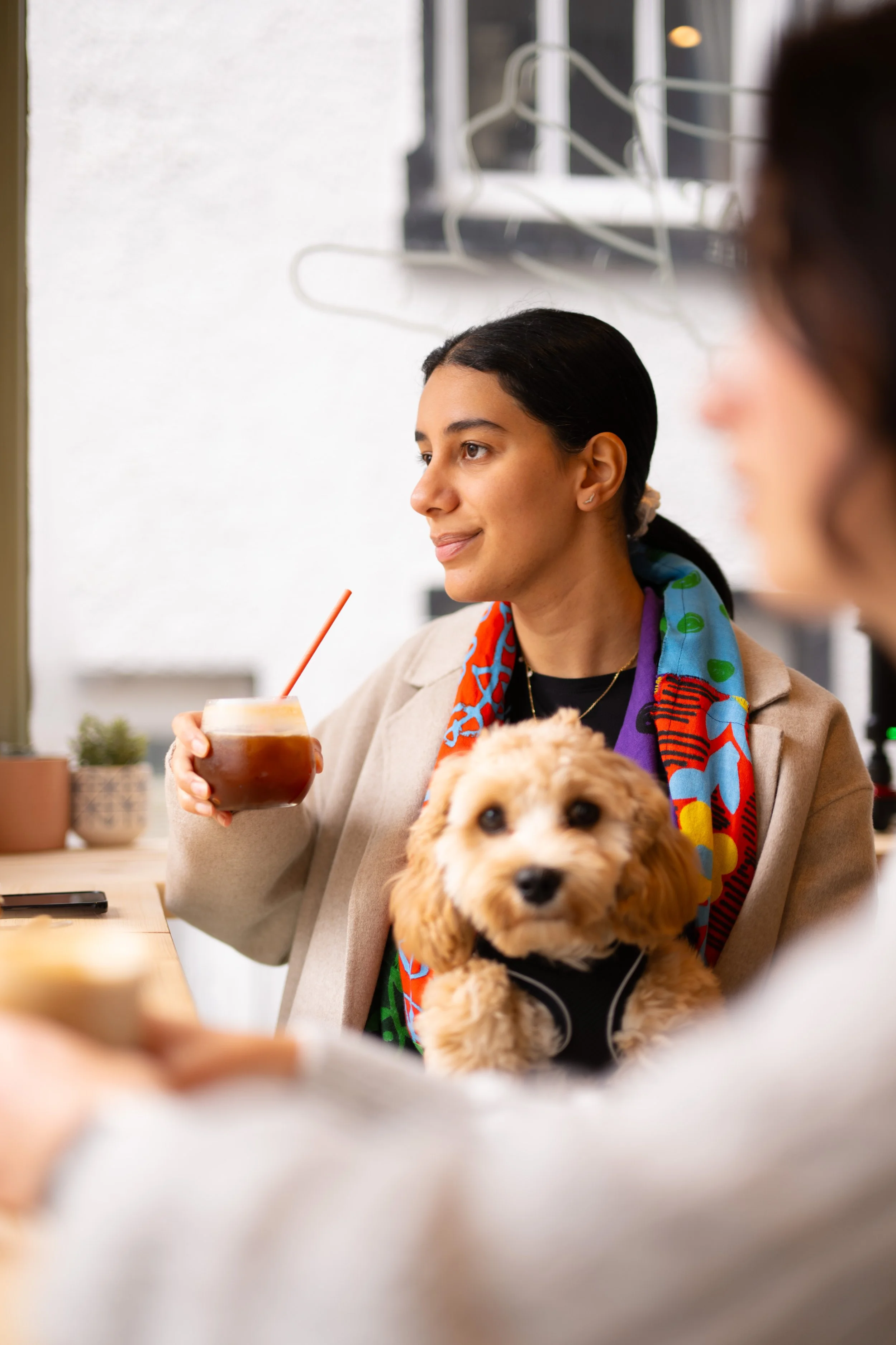 A woman with dark hair tied back, wearing a colorful scarf and beige coat, is sitting at a table holding a glass of cold beverage with a straw. She has a small, fluffy, light brown puppy with dark eyes and nose sitting on her lap. There are blurred hands in the foreground, possibly of other people in the cafe, and a person with glasses and dark hair is partially visible on the right. The background shows a white wall with a wall-mounted black chalkboard and some white wire hangers.