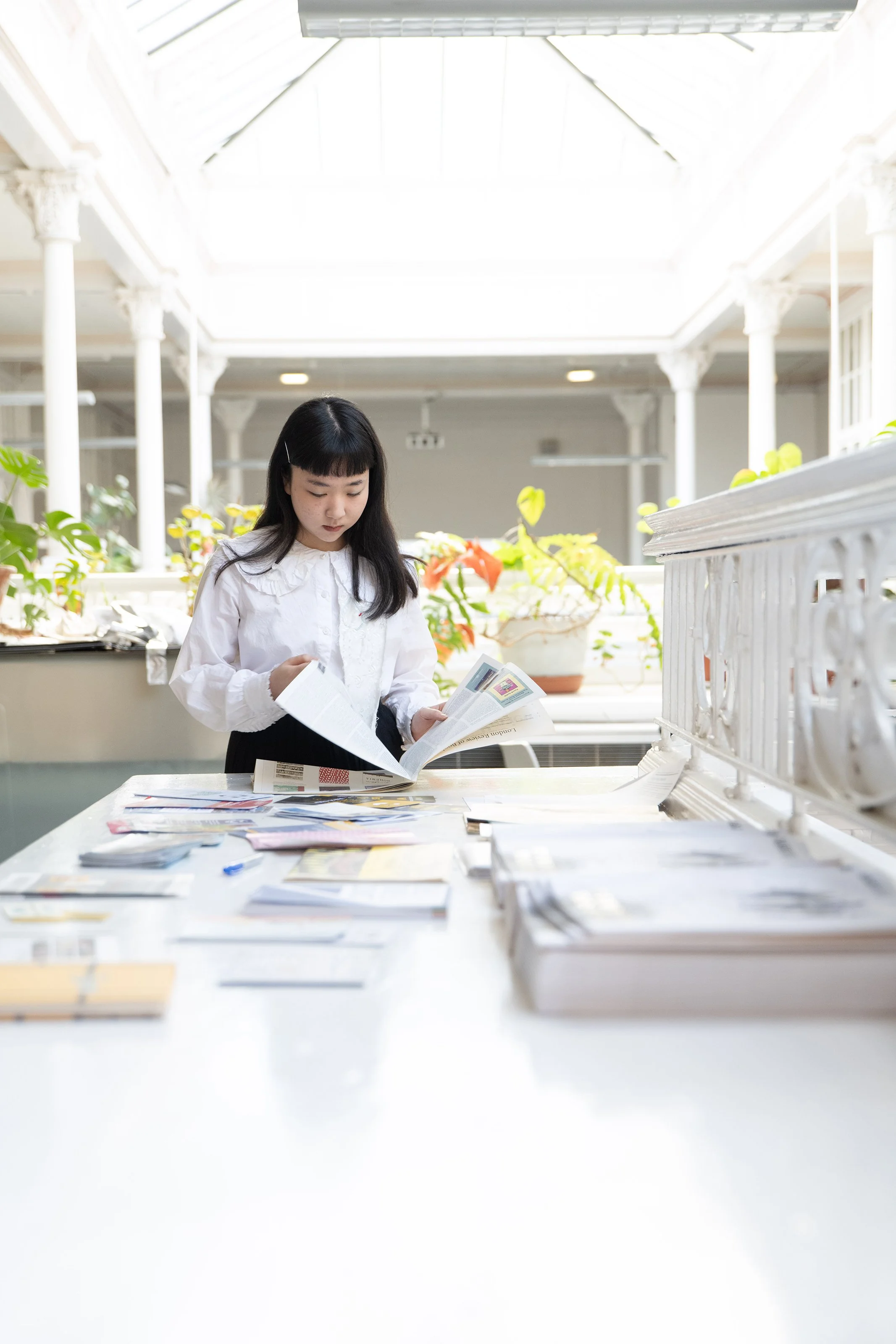 A young woman with long black hair and bangs, wearing a white blouse, stands at a table with magazines and papers, reading a booklet or brochure in a bright, airy space with high ceilings, white columns, and potted plants.
