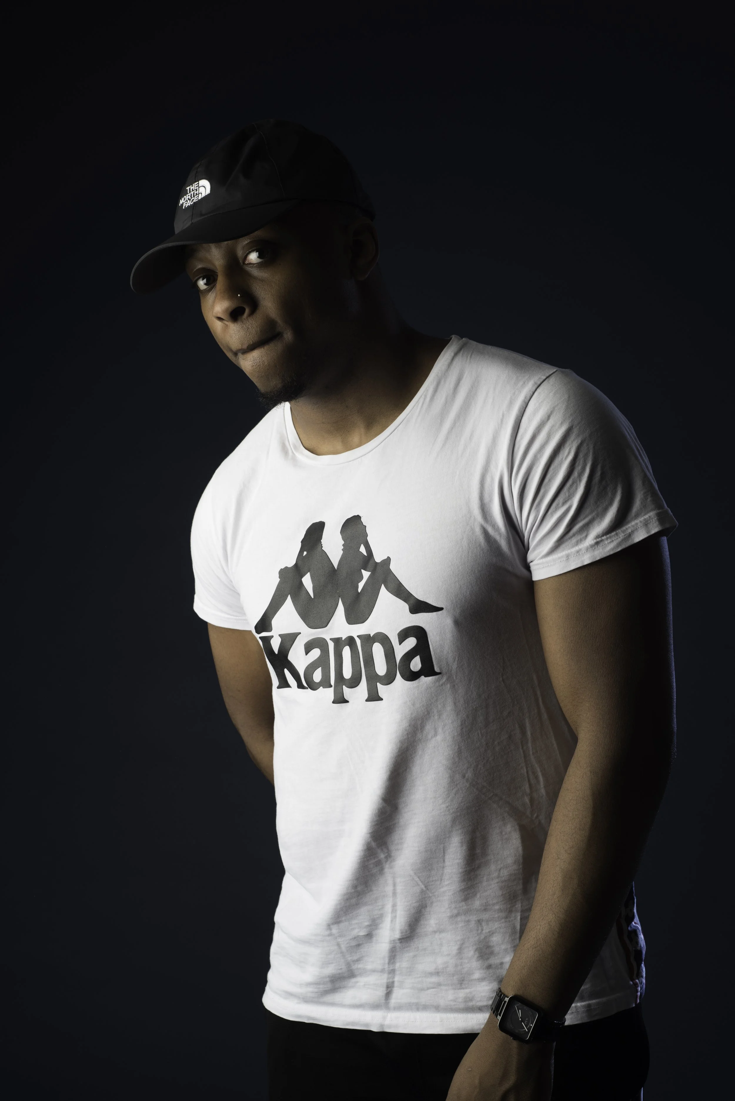 A young man in a white Kappa t-shirt with a graphic logo, wearing a black North Face cap, and a black watch on his left wrist, standing against a dark background.