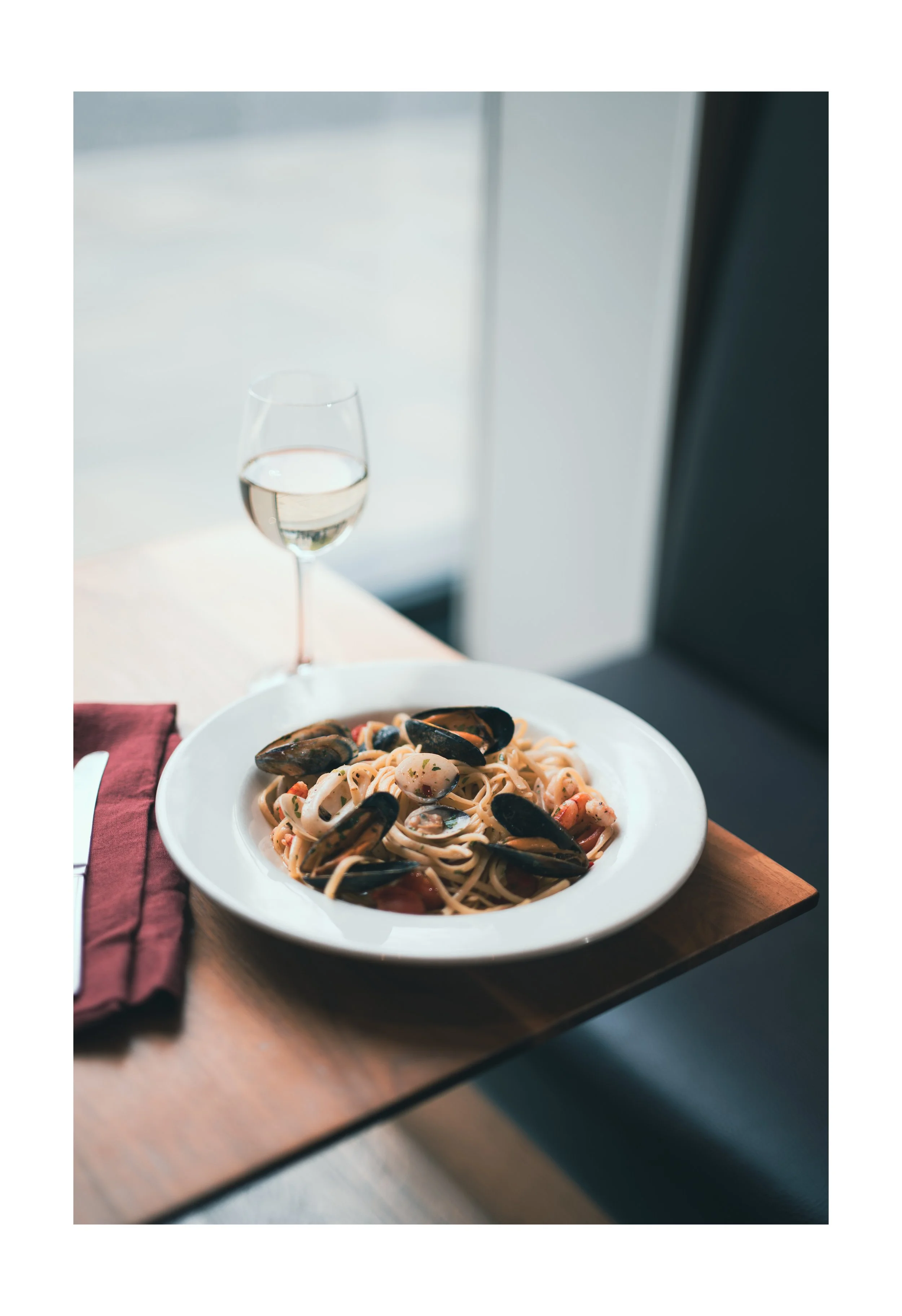 A plate of seafood pasta with mussels, shrimp, and pasta on a wooden tray with a glass of white wine in the background near a window.