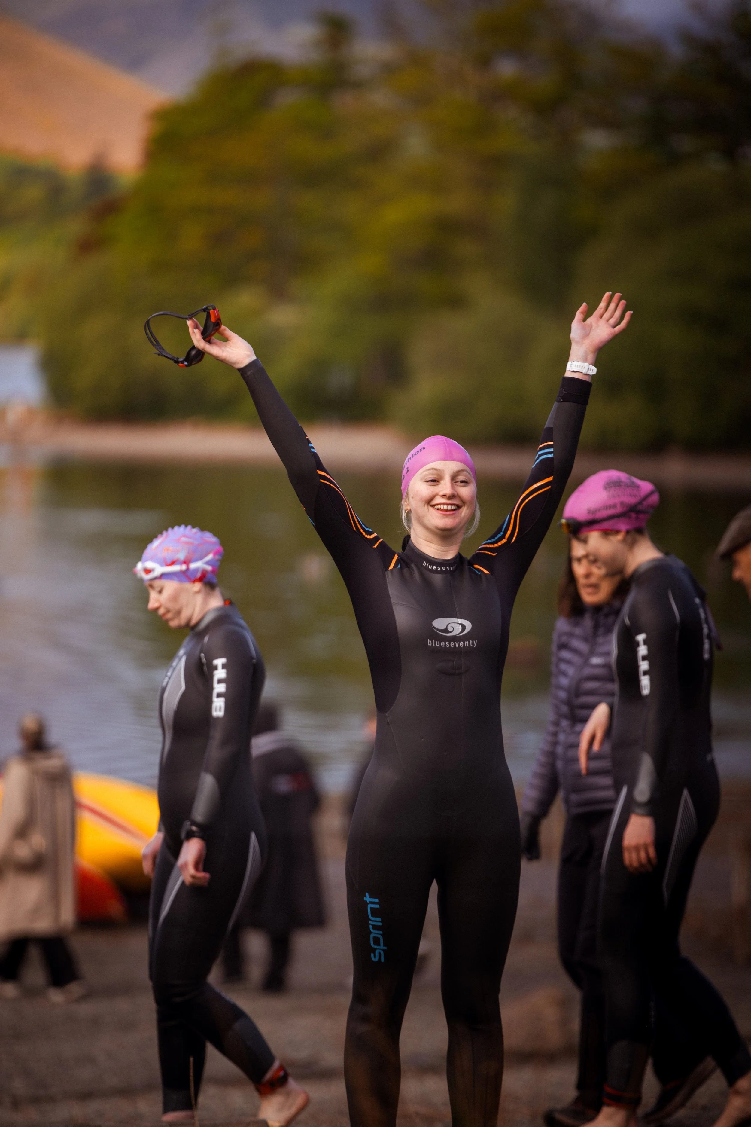 A group of people wearing wetsuits by a river, with one woman raising her arms and smiling, holding goggles in her right hand during a sporting event.