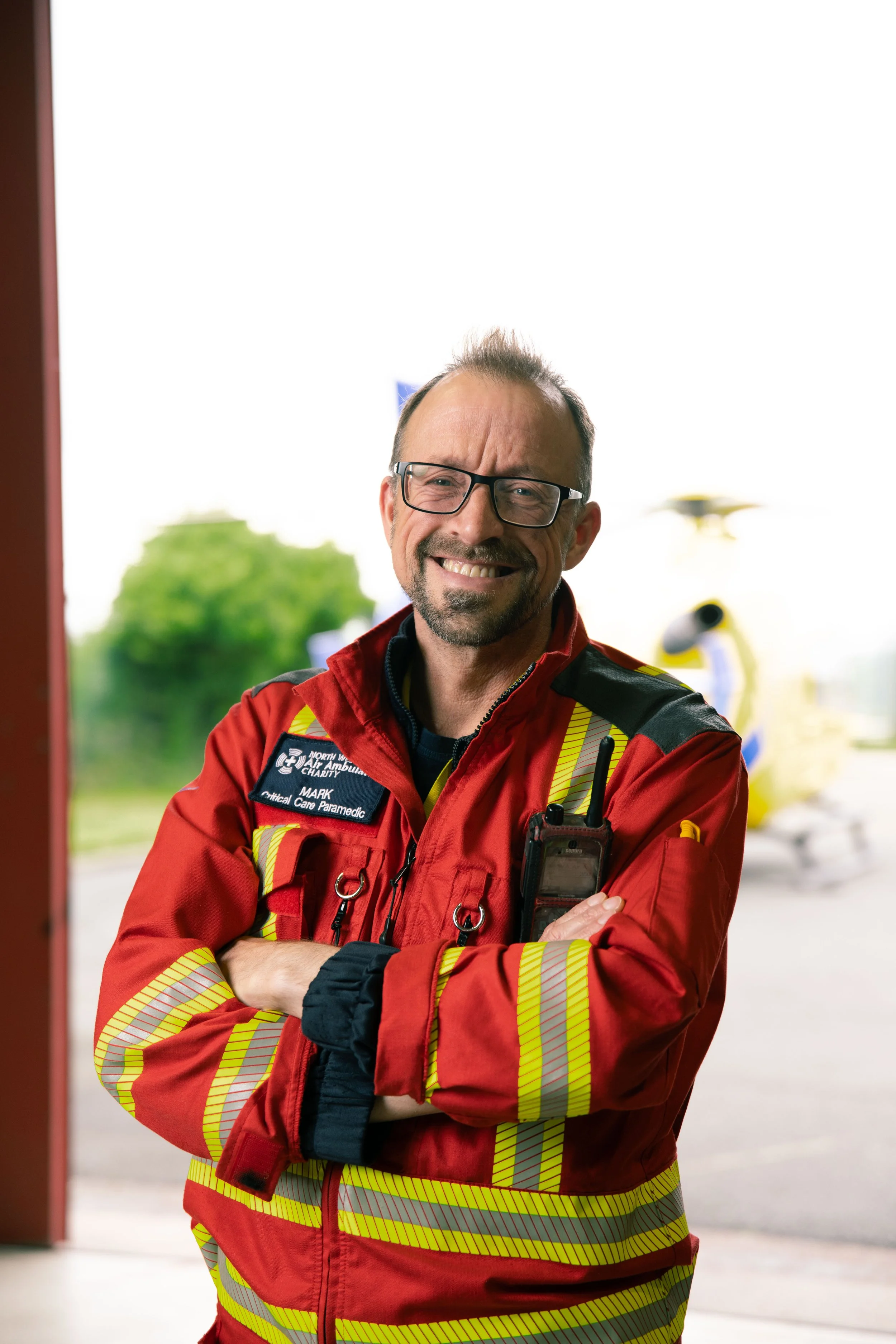 Smiling male paramedic in red and yellow uniform with glasses, standing with arms crossed.