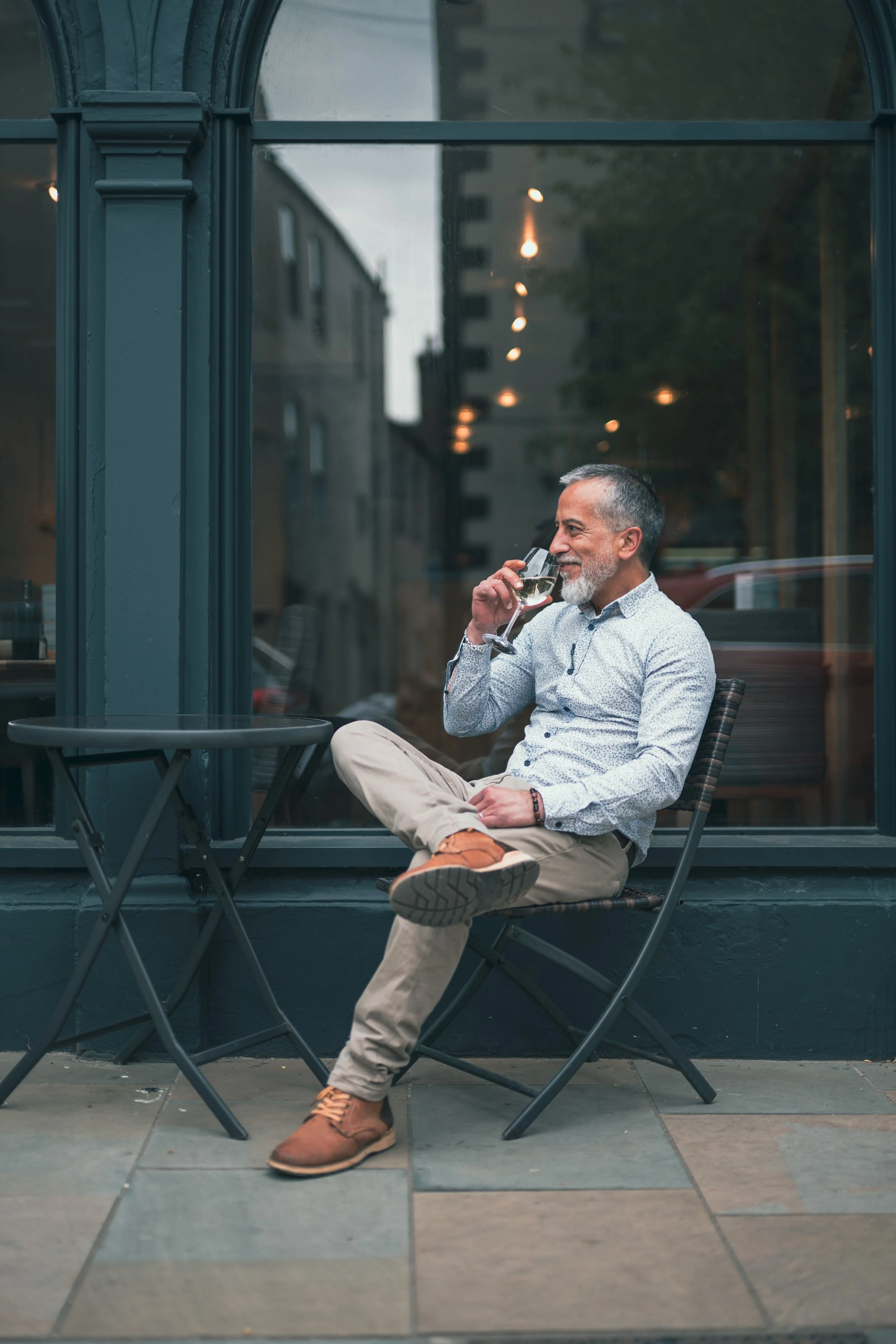 A middle-aged man with gray hair and beard, wearing a light-colored patterned shirt and beige pants, sitting outside at a cafe with his right leg crossed over his left knee, smiling, and drinking white wine from a glass.