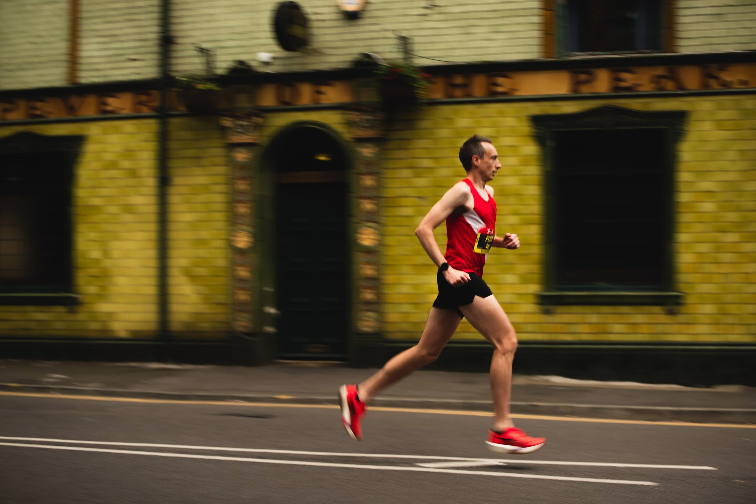 A man running outdoors on a street, wearing a red sleeveless shirt, black shorts, and red running shoes, with a yellow race bib attached to his shirt.