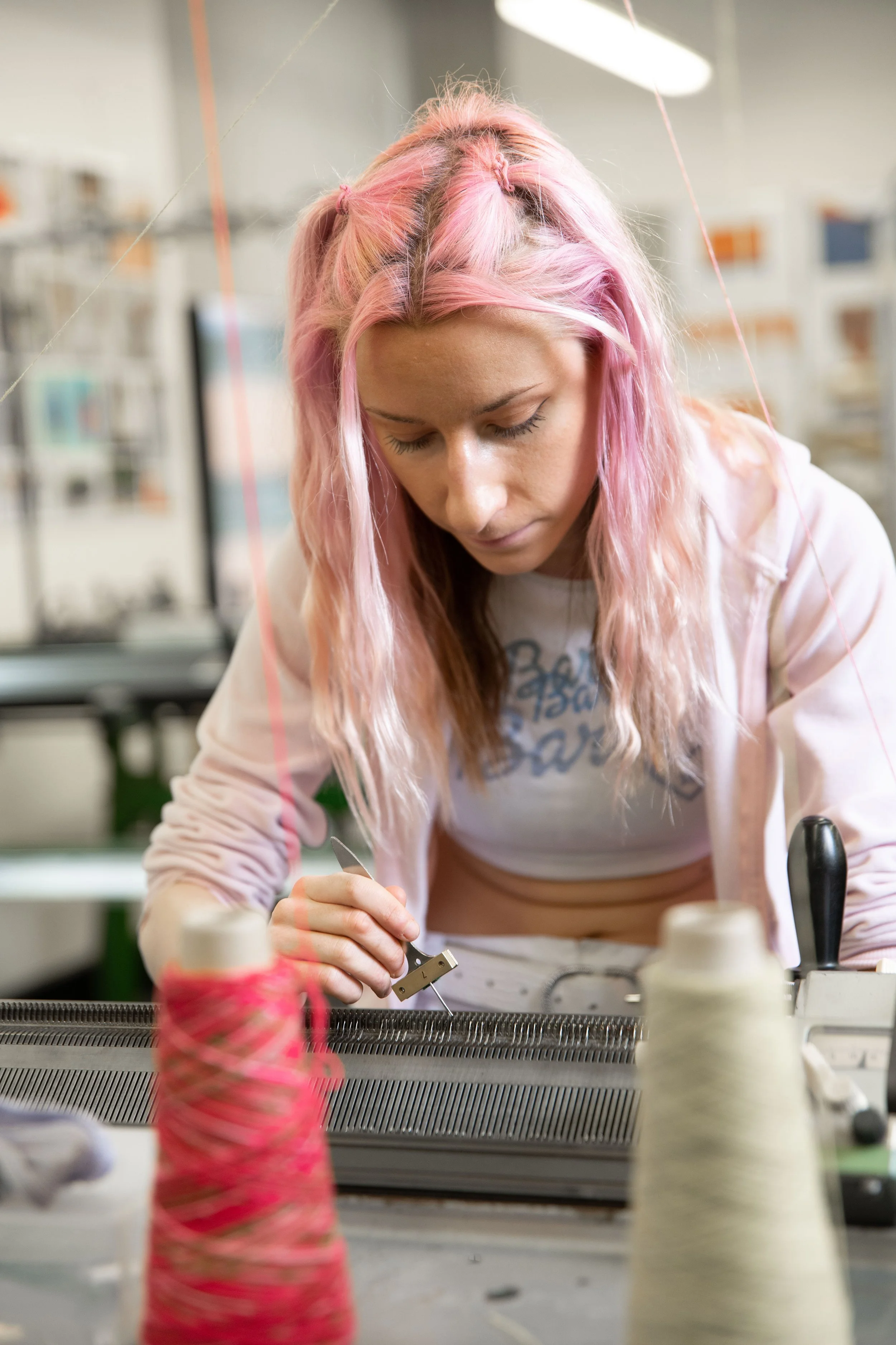 A woman with pink hair in pigtails working with a tool at a table, surrounded by spools of yarn and yarn in a workshop or craft room.