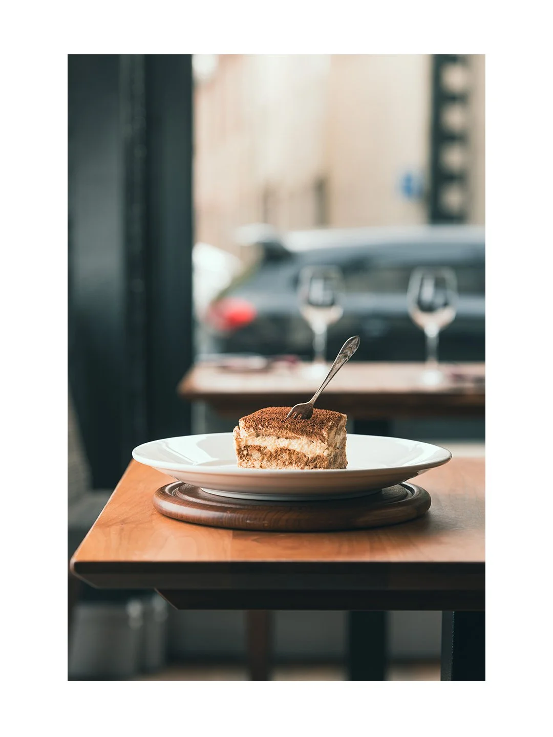 A slice of tiramisu on a white plate with a fork, placed on a wooden table in a cafe with a blurry car and buildings outside the window.