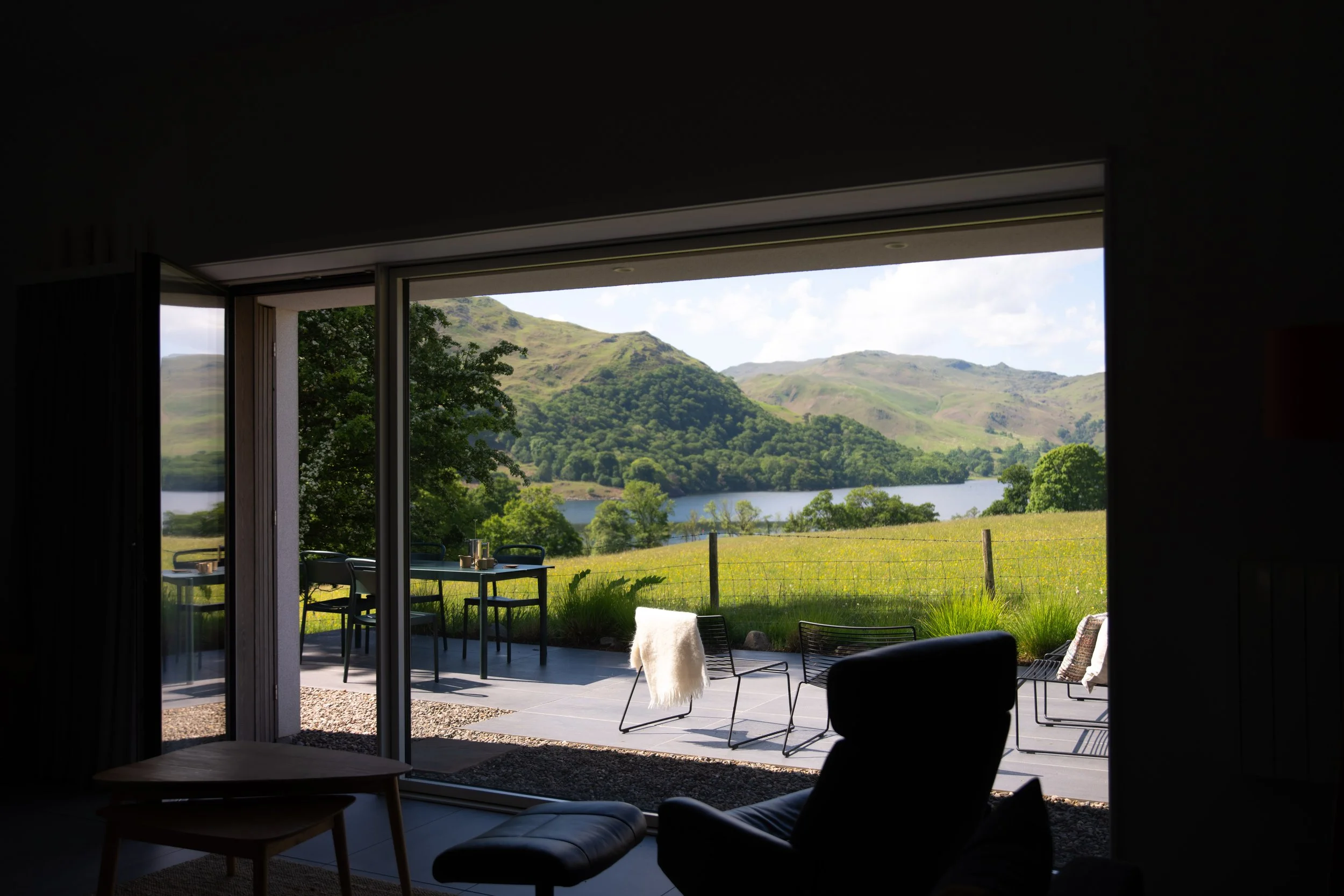 Interior view looking out through open sliding glass doors to a patio with outdoor furniture, surrounded by lush green hills and a lake in the distance under a partly cloudy sky.