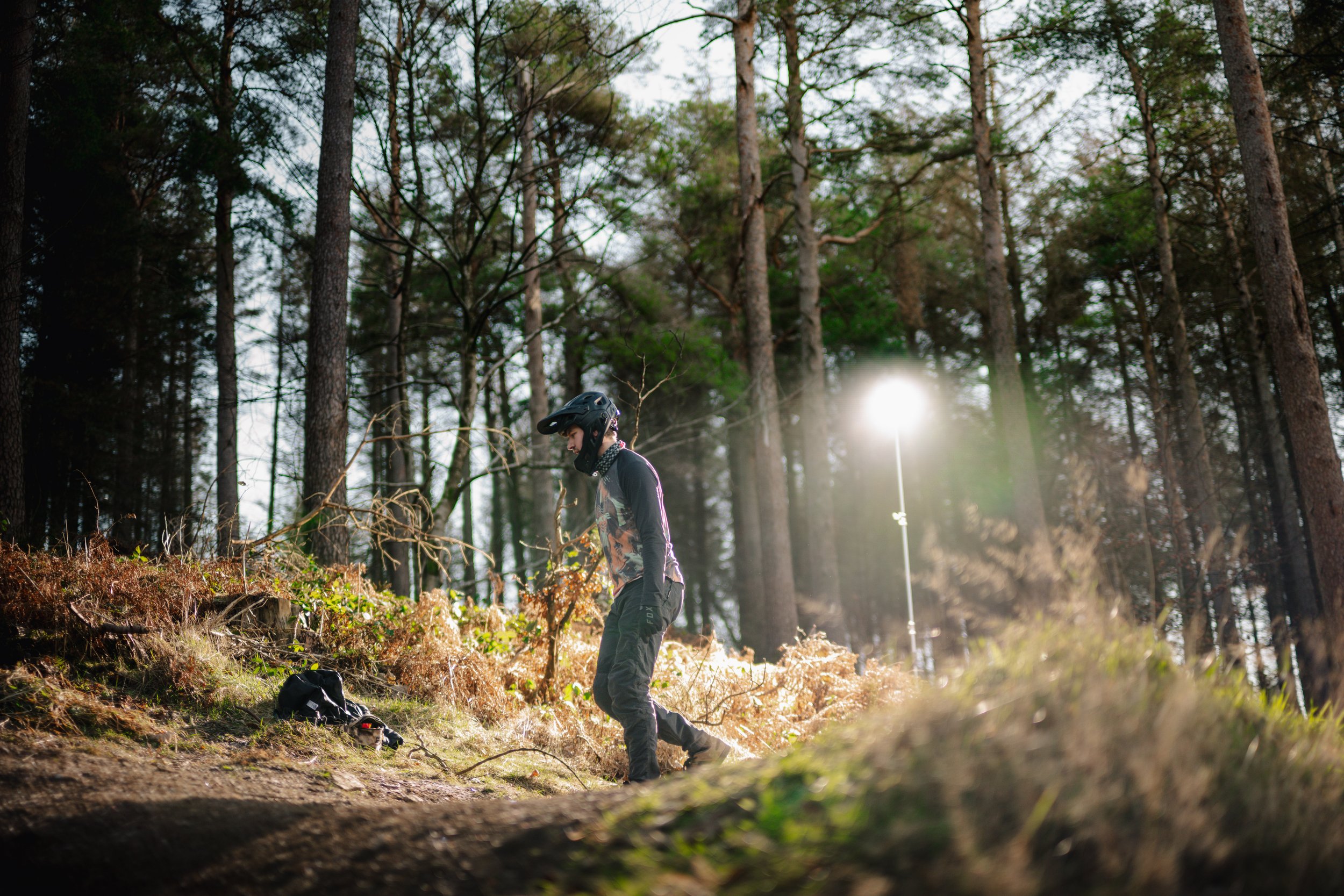 A person wearing a helmet and outdoor clothing is standing in a wooded area with tall trees, preparing for an outdoor activity during sunlight.