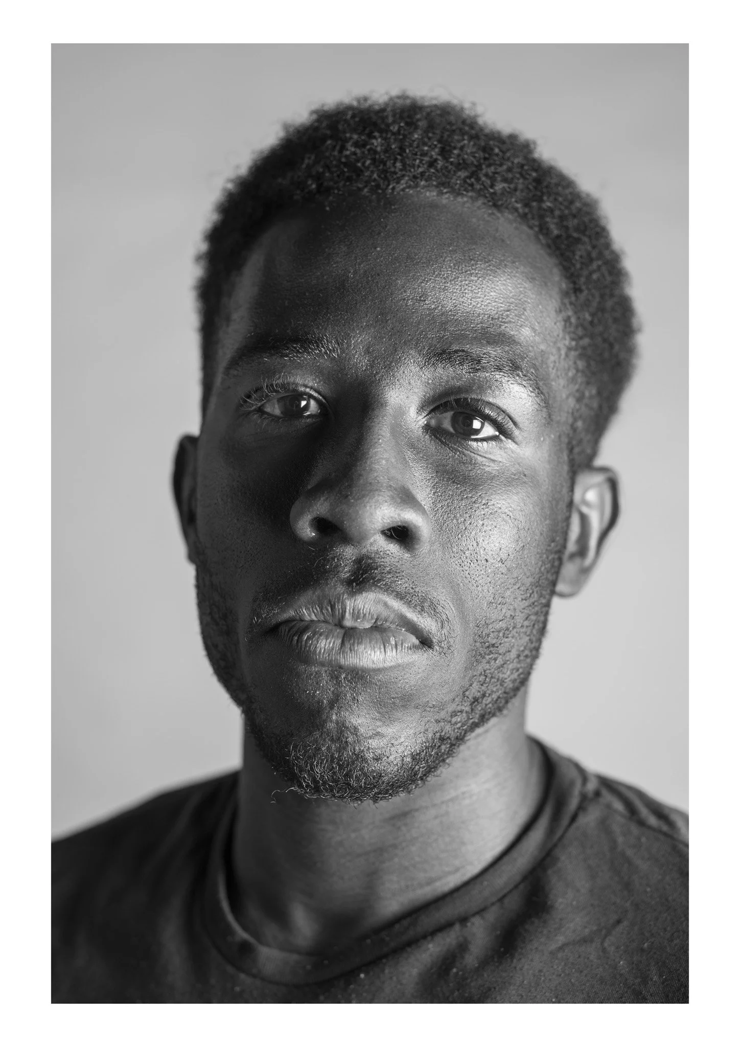 Black and white portrait of a young man with short curly hair, smooth skin, and a slight beard, wearing a dark t-shirt, looking directly at the camera.