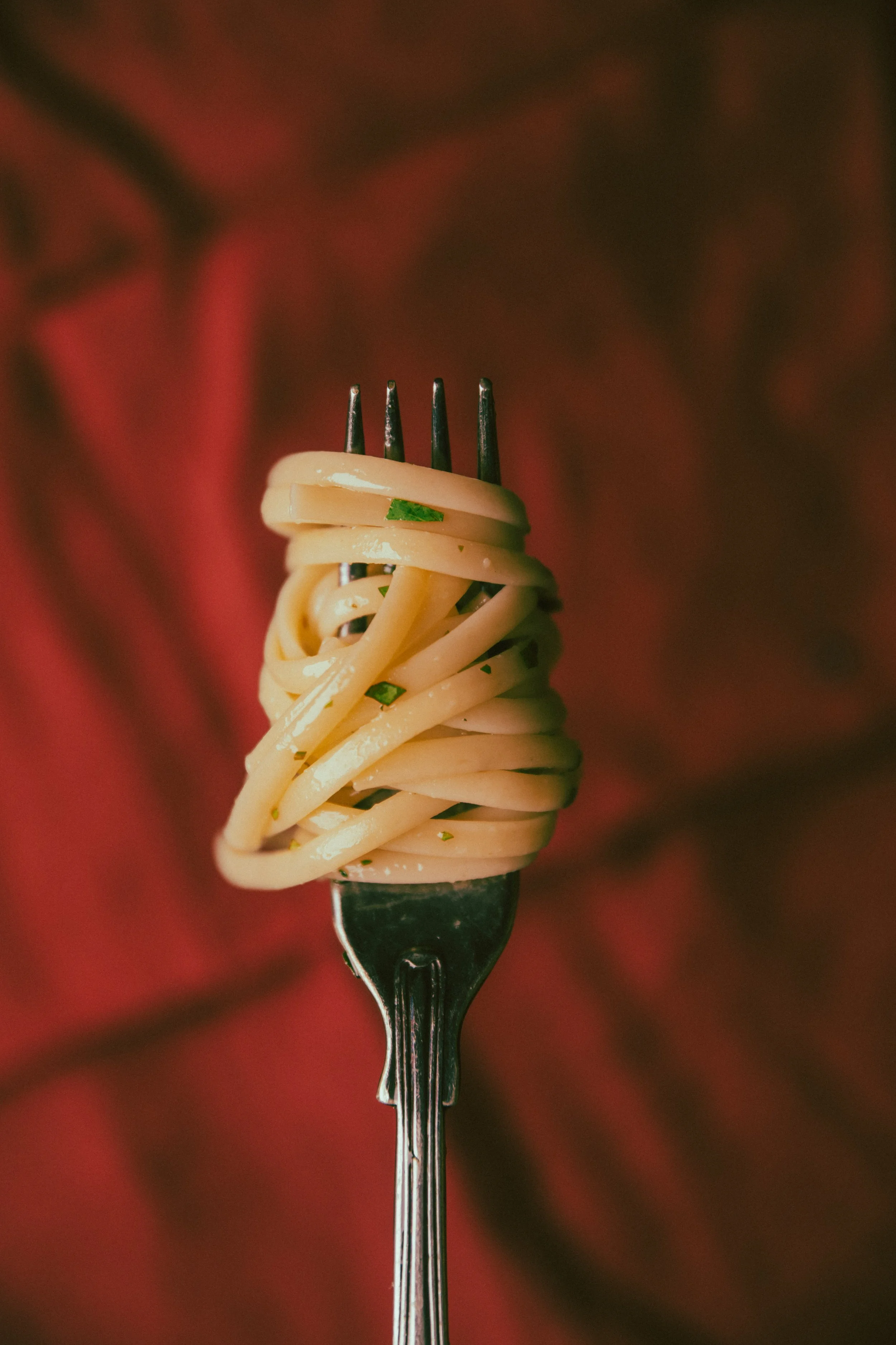 A fork with a twisted nest of cooked spaghetti noodles garnished with chopped herbs, against a blurred dark background.