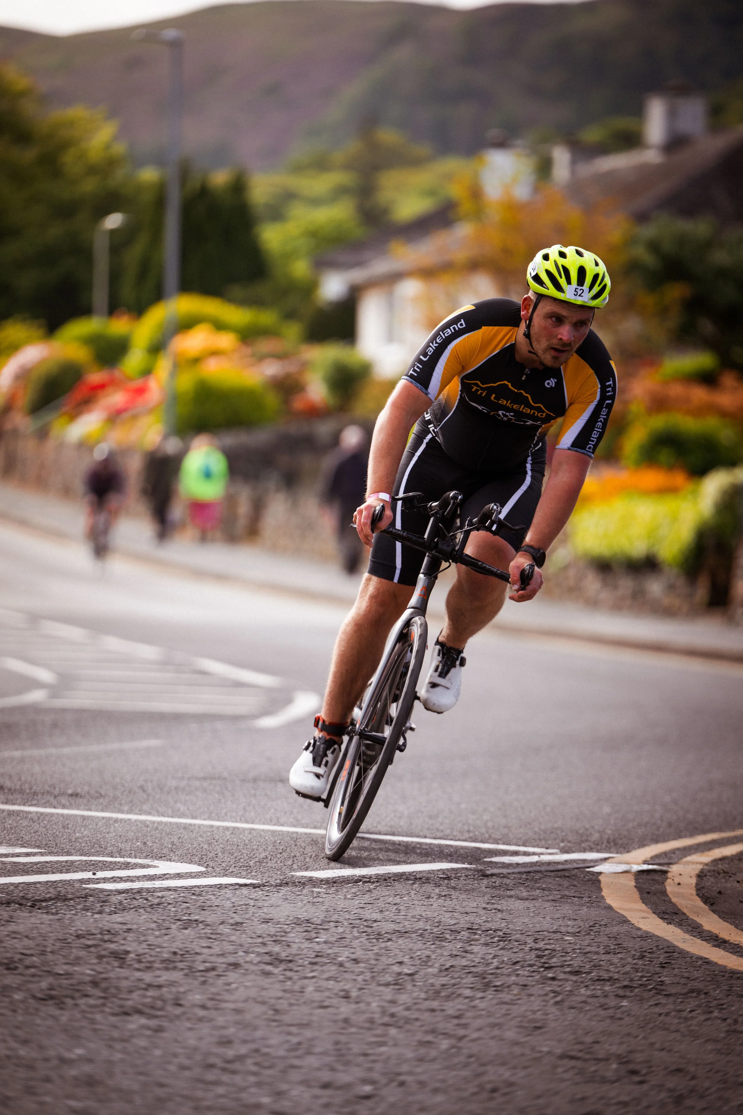 A cyclist with a yellow helmet and black and yellow cycling jersey leaning into a turn on a road in a residential area with houses and autumn-colored trees in the background.