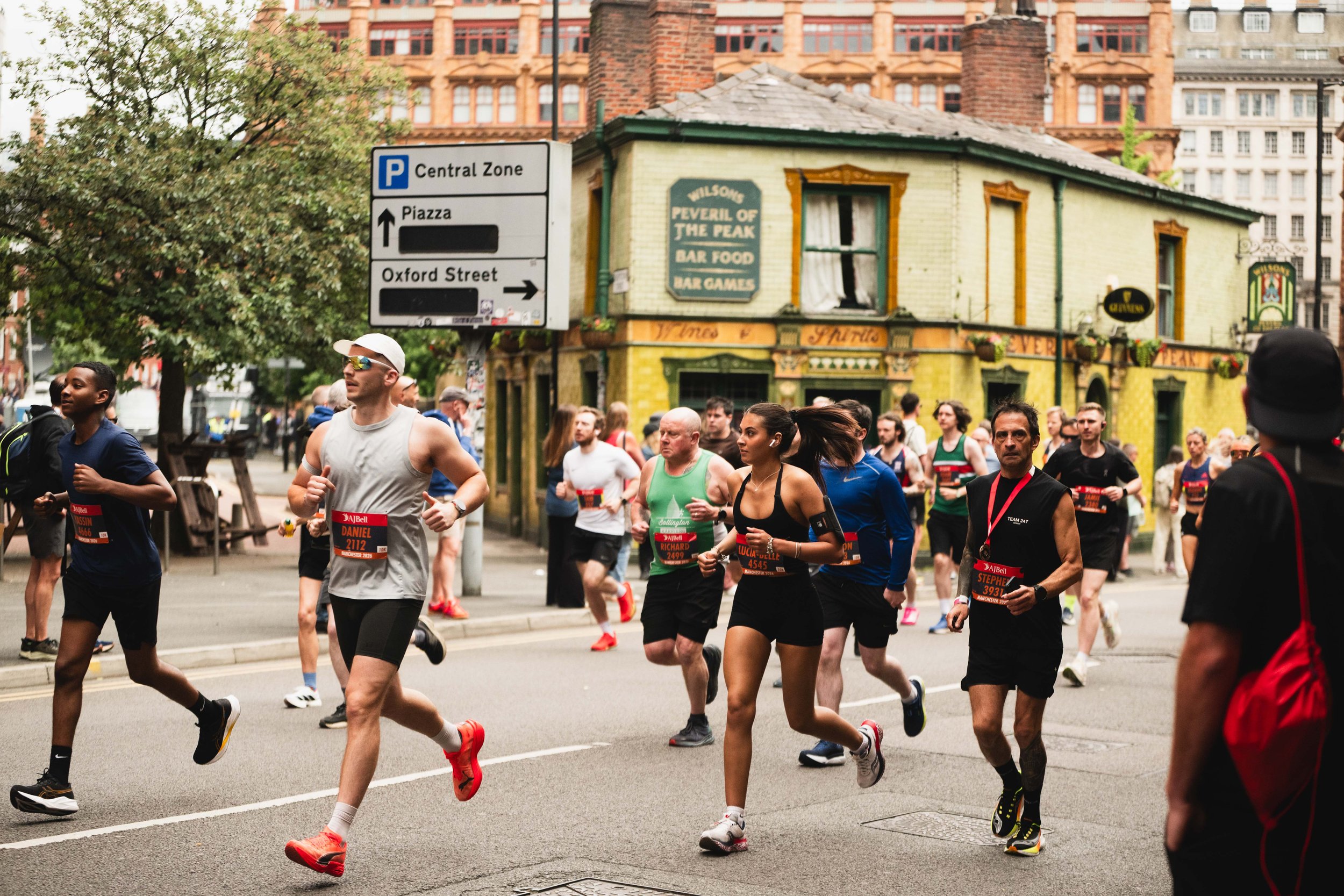 A group of marathon runners participating in a race on a city street. Some runners are wearing bibs with numbers, and one man is wearing sunglasses and a white cap. The background features a historic building and a street sign indicating parking, Pia