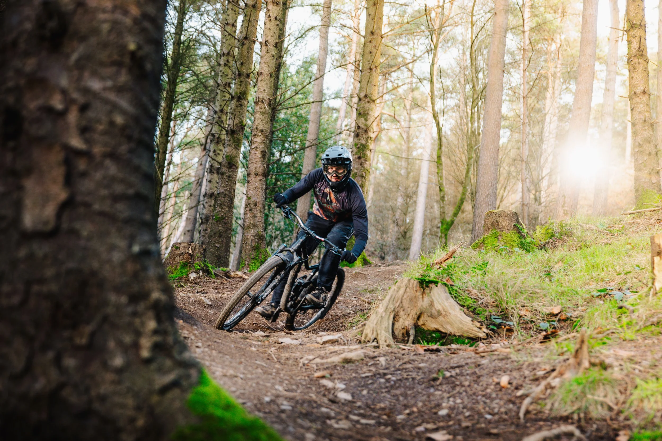 A person wearing a helmet and glasses mountain biking on a trail through a forest with tall trees and sunlight filtering through the branches.