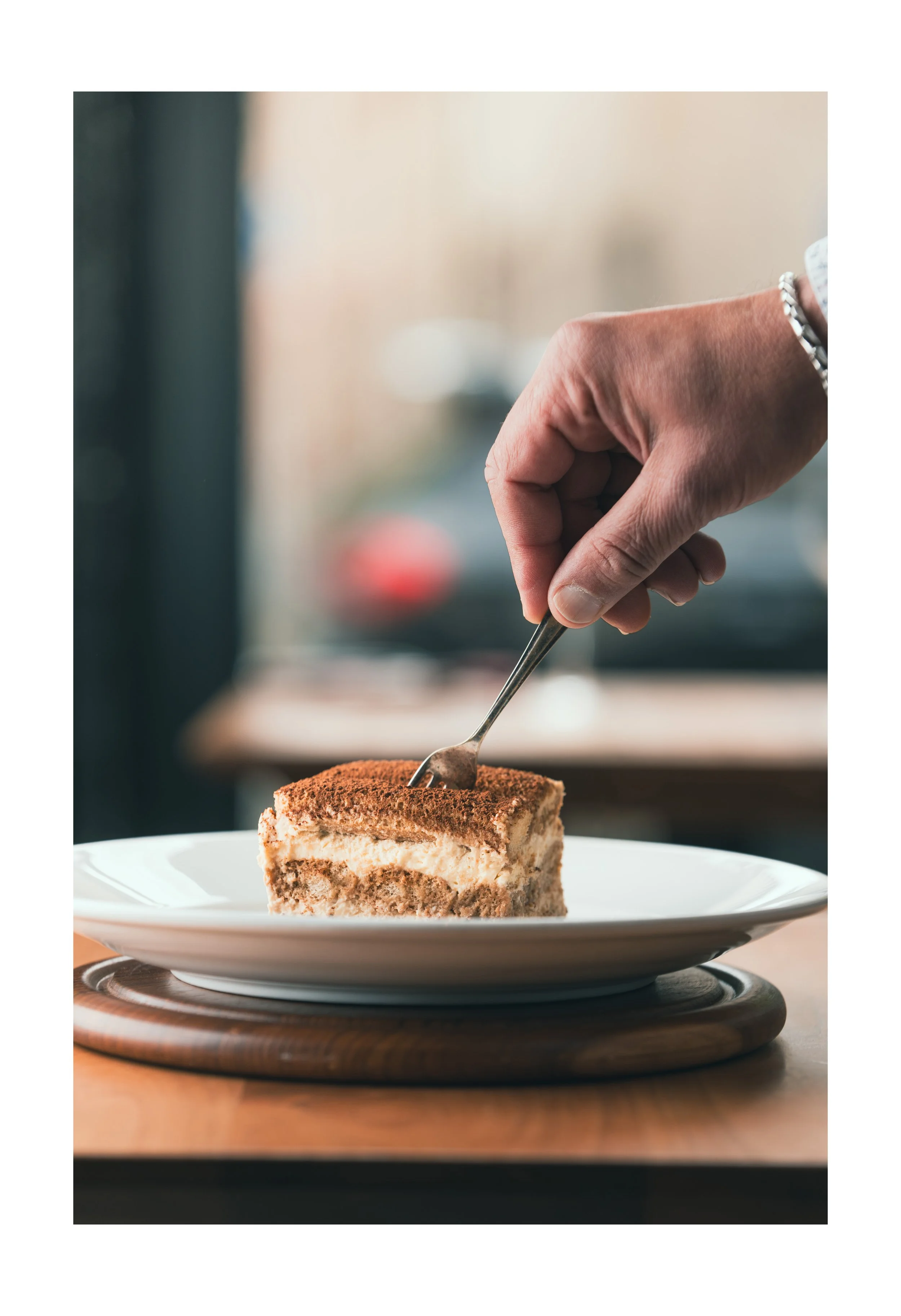 Close-up of a hand using a fork to serve a slice of tiramisu cake on a white plate.