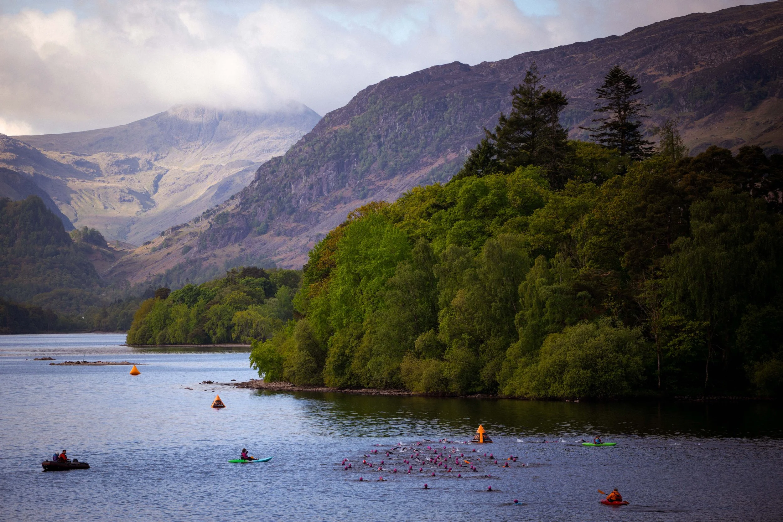 A landscape of a lake surrounded by green trees, with mountains in the background, some partially covered by clouds. There are swimmers in the water with bright pink caps, and a few people in kayaks or on boats near the shore. Orange buoys mark the swimming area.