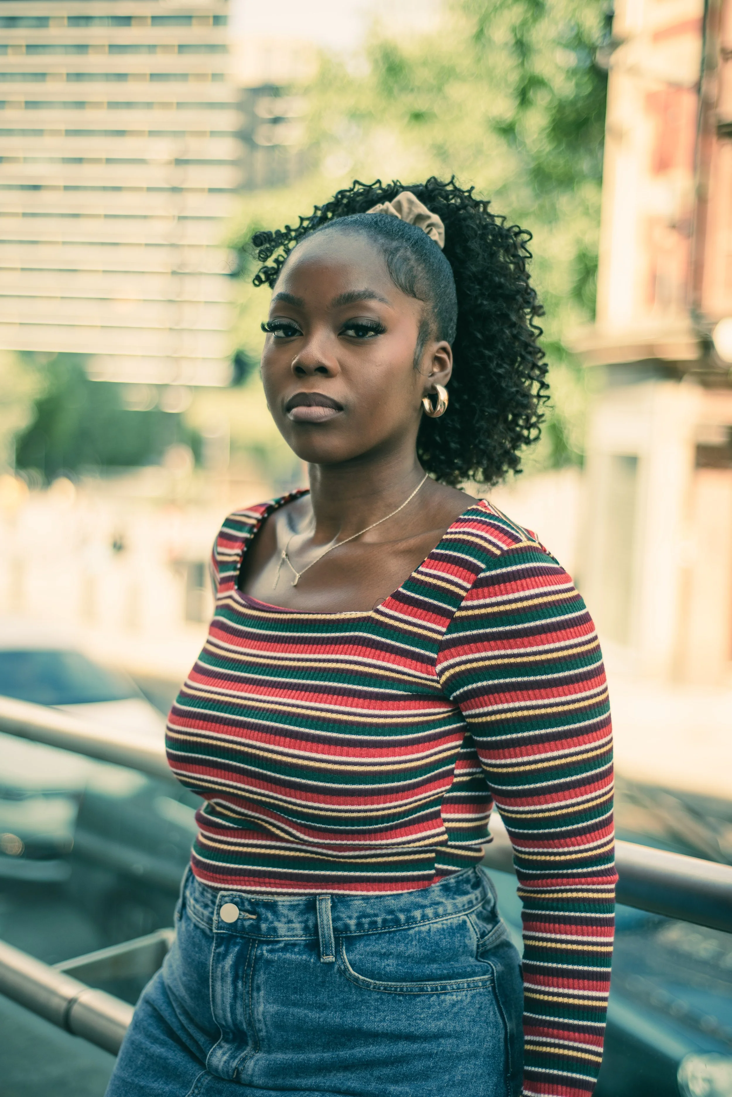 Portrait of a young woman with curly hair tied up with a beige scrunchie, wearing gold hoop earrings, a colorful striped long-sleeve shirt, and high-waisted jeans, standing outdoors near a railing with a blurred cityscape and greenery in the backgrou