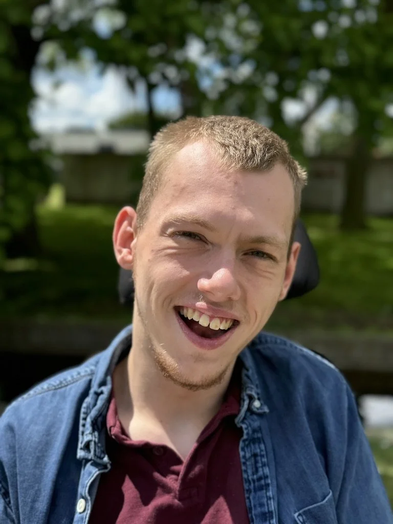 A young man with short blonde hair and a goatee smiling outdoors, wearing a maroon shirt and denim jacket, with trees and blue sky in the background.