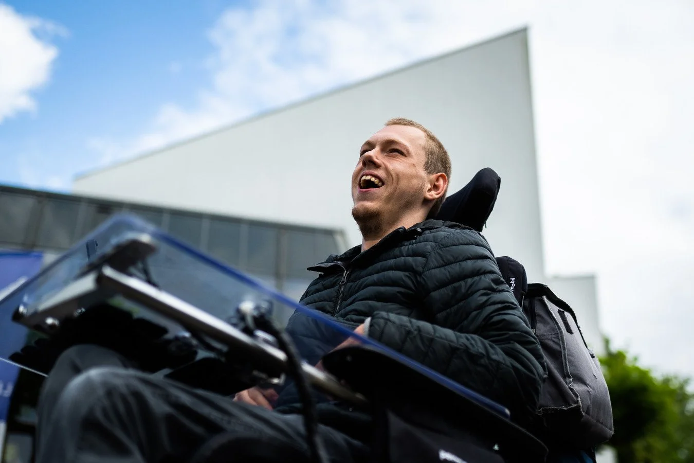 Young man with a backpack sitting outdoors, smiling and looking upward on a cloudy day.