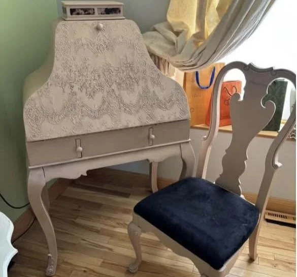 A vintage-style writing desk with intricate patterns, paired with a matching chair with a dark blue cushion, placed near a window with curtains and a glimpse of an orange bag outside.