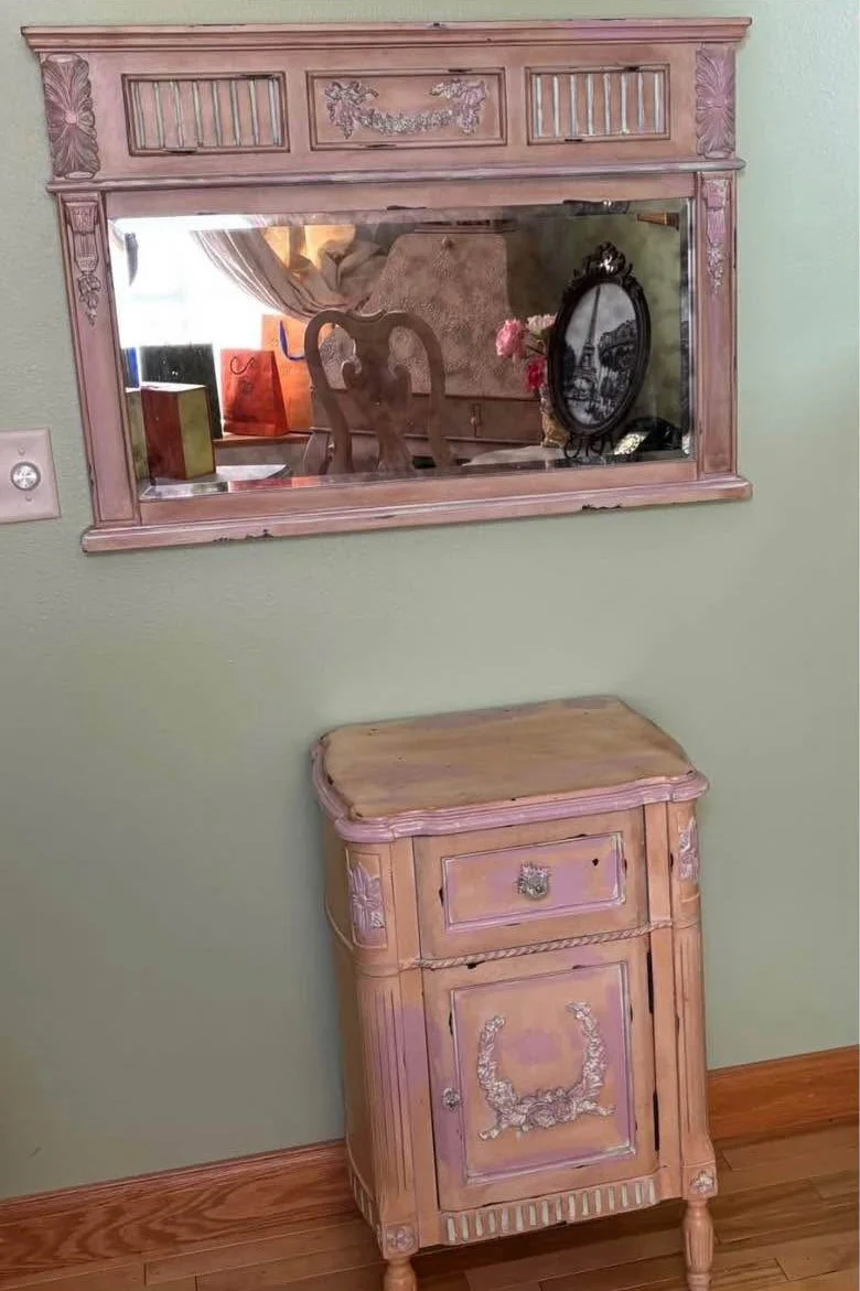 A vintage wooden mirror with ornate pinkish finish and carved decorative details hangs above a matching small wooden cabinet with a single drawer and door, both on a green wall. The mirror reflects a room with a chair, curtain, and various objects.