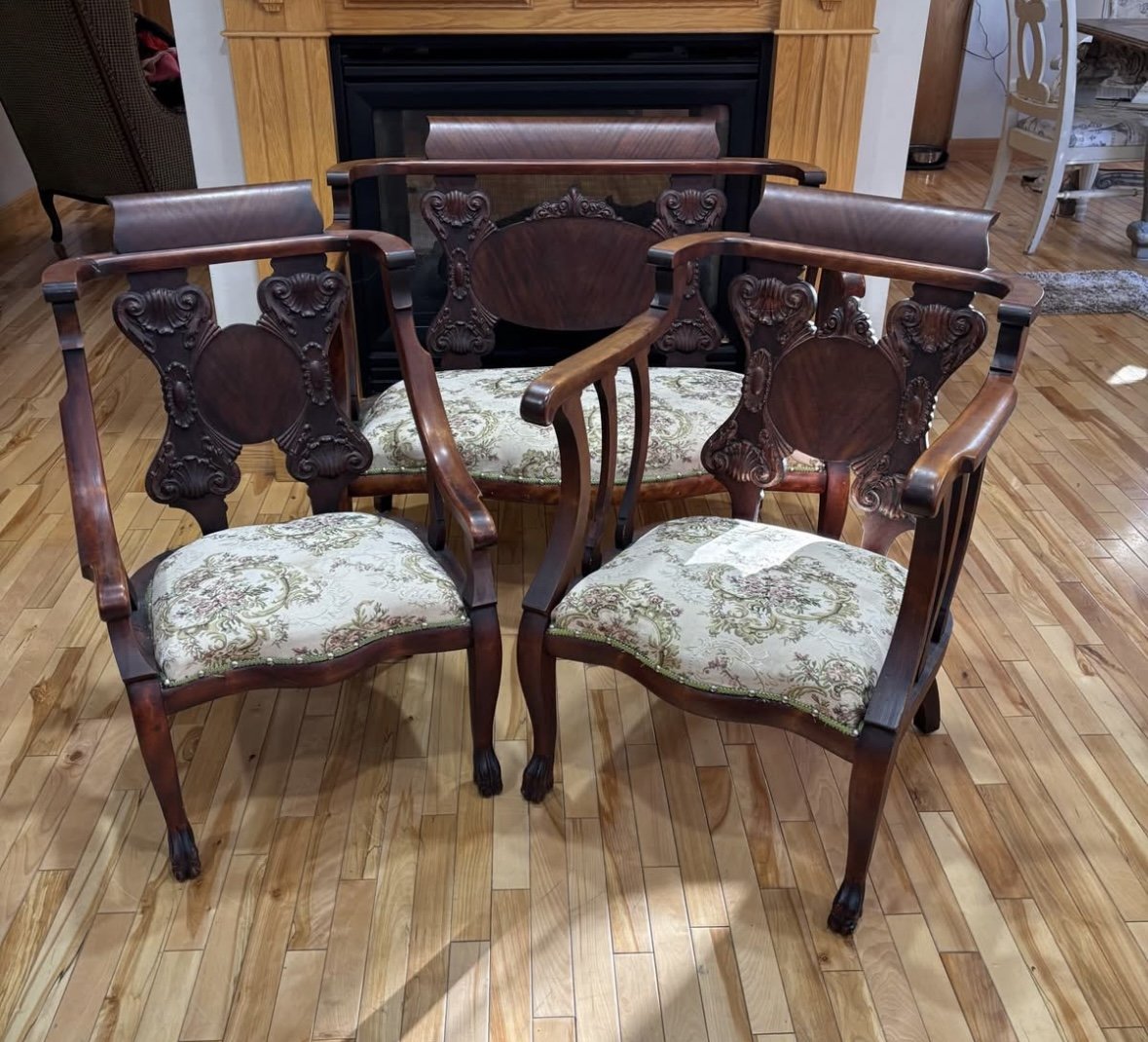Set of four vintage wooden chairs with upholstered seats and ornate carved details, placed on a hardwood floor in front of a fireplace.