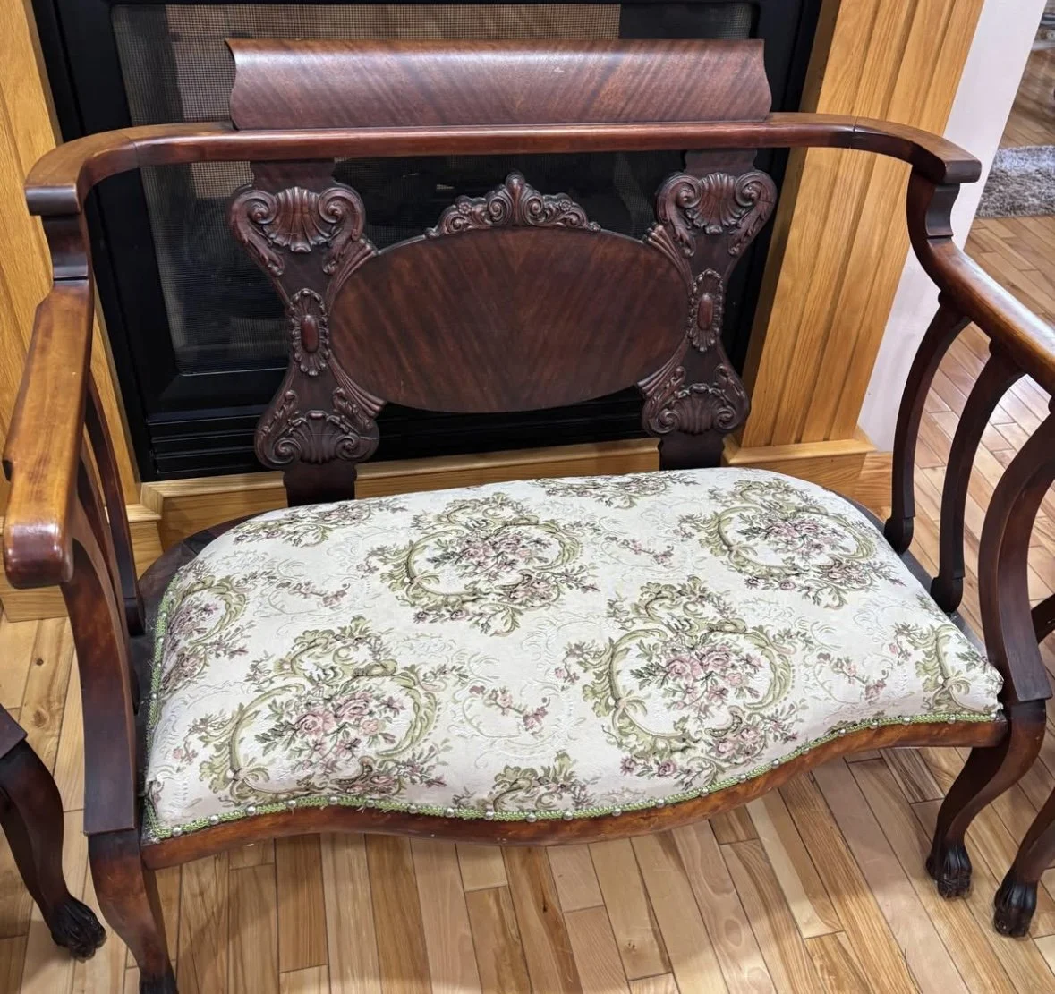 A vintage wooden bench with carved backrest and floral cushioned seat, placed in front of a fireplace with a wooden surround.