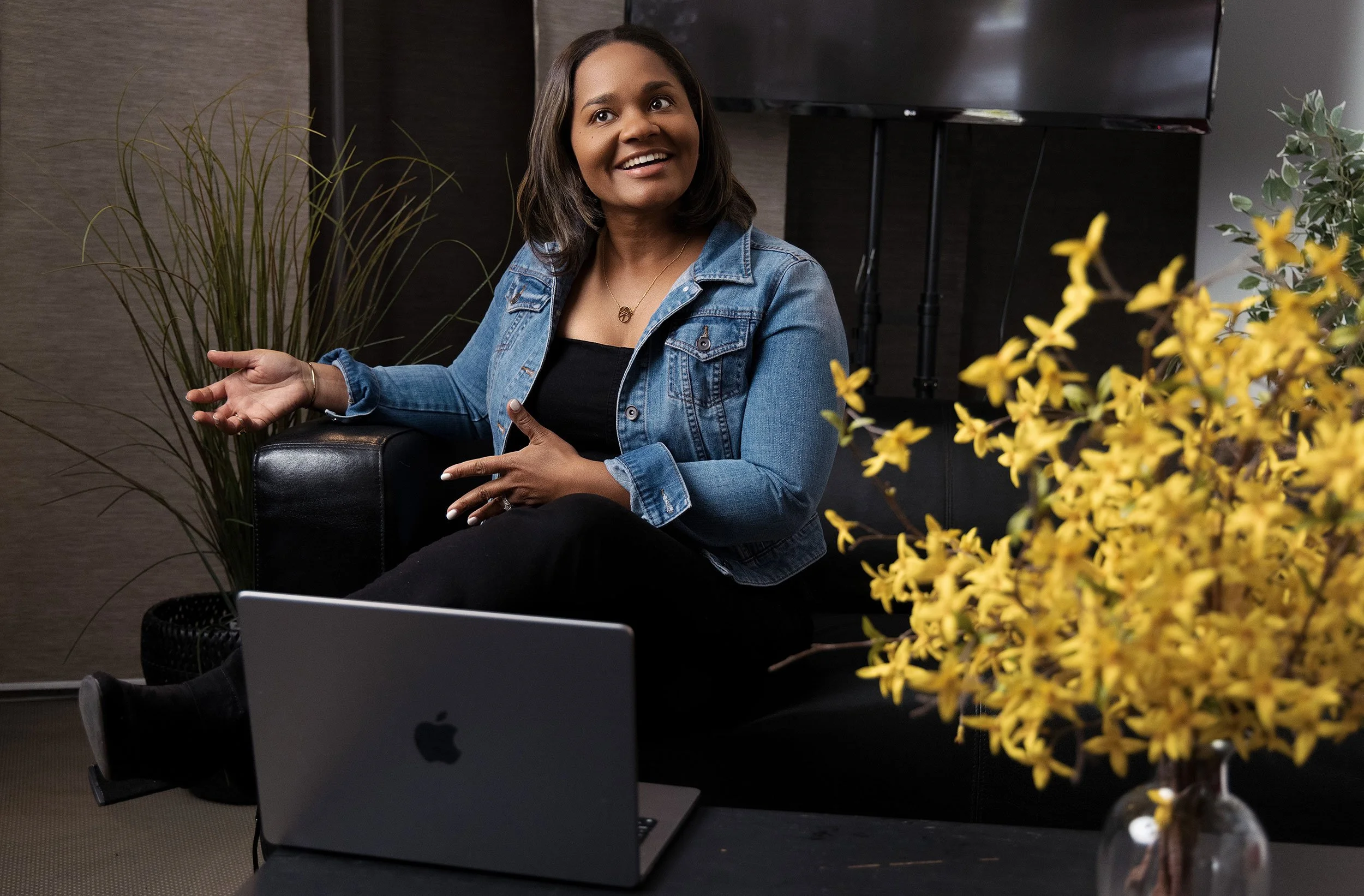 A female Delaware life coach wearing a denim jacket, flanked by a yellow plant uses hand gestures while speaking to a client