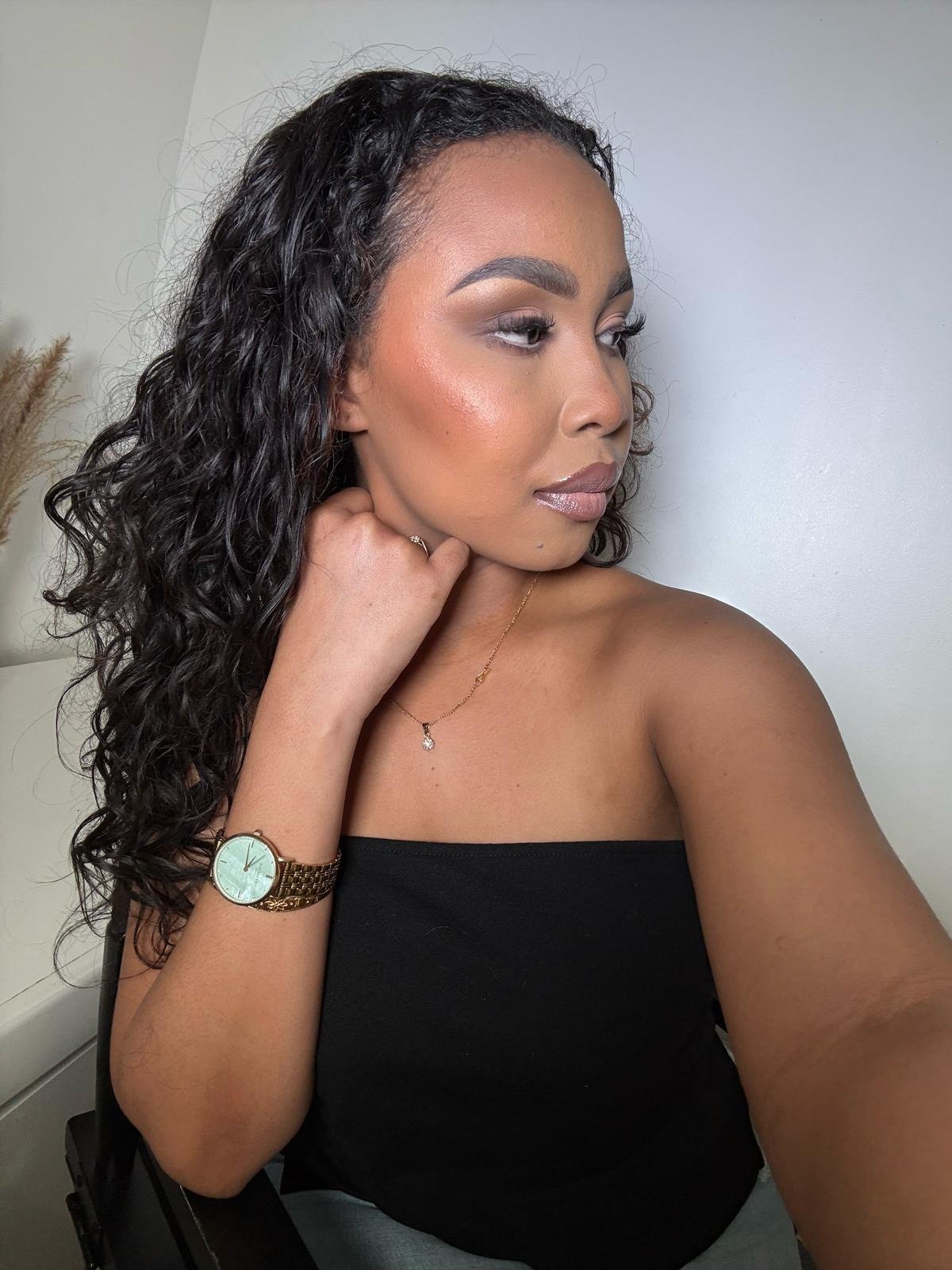 Femme avec cheveux bouclés noirs, portant une montre dorée et un collier, vêtue d'un haut noir, posant avec un fond clair.