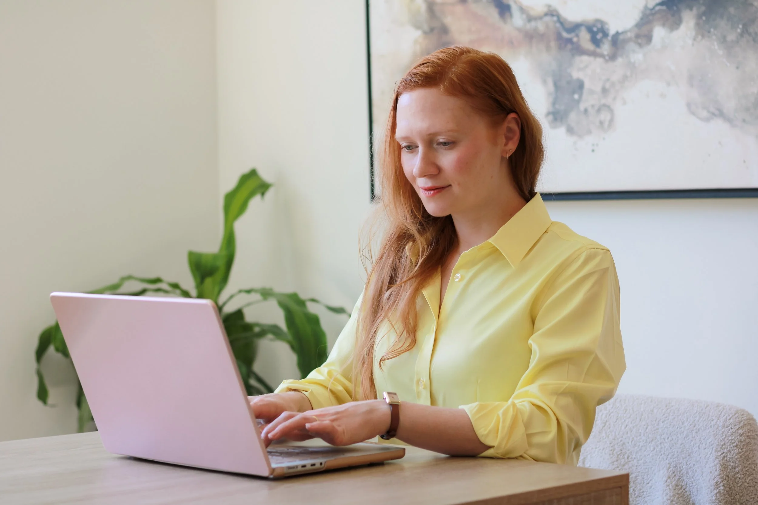 A woman with red hair in a yellow blouse sitting at a wooden table working on a silver laptop in an office, with a large abstract painting on the wall behind her and a plant in the background.