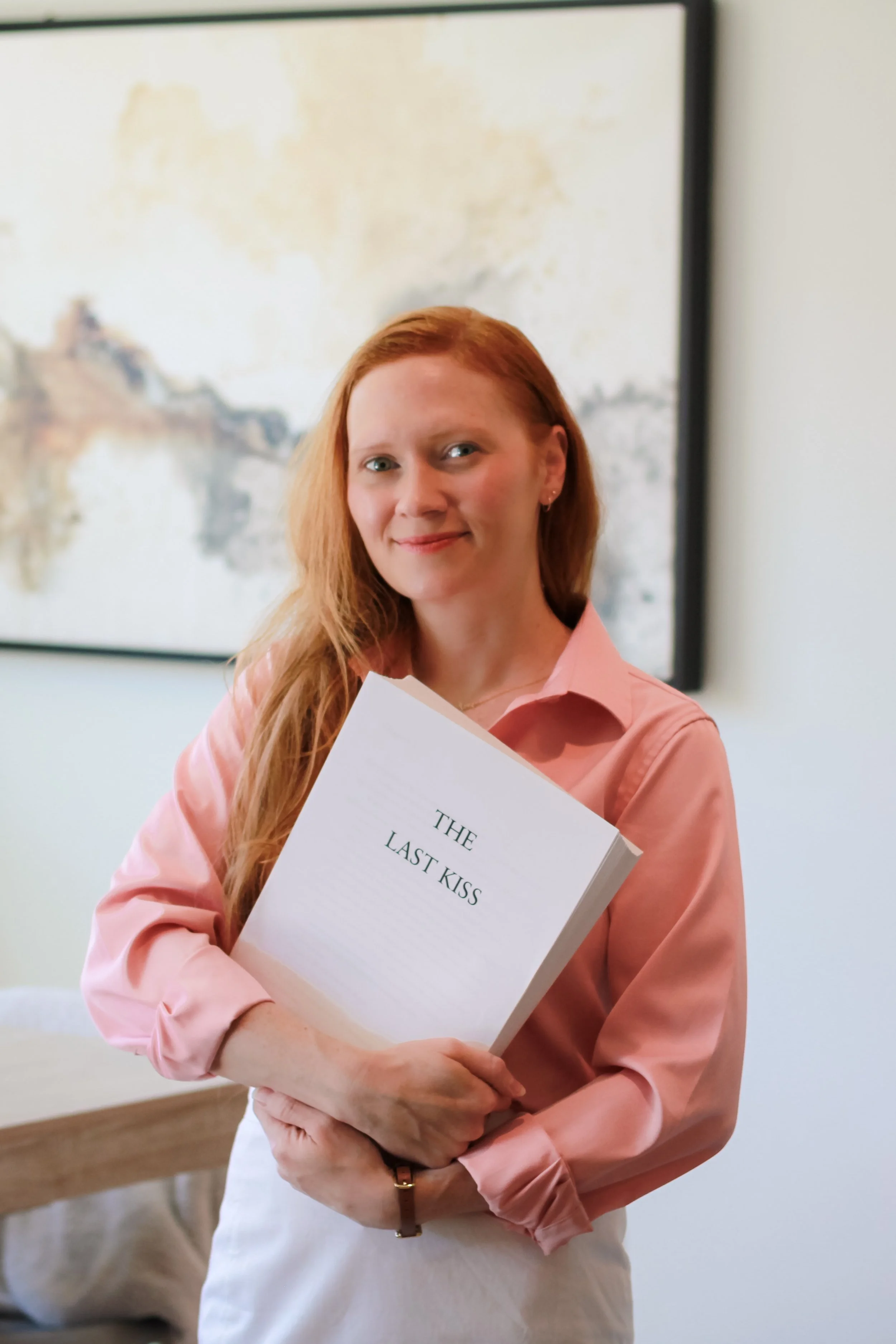 A woman with long red hair, wearing a pink blouse, is holding a white book titled "The Last Kiss." She is smiling and standing in front of a blurred background with light-colored walls and a large abstract painting.