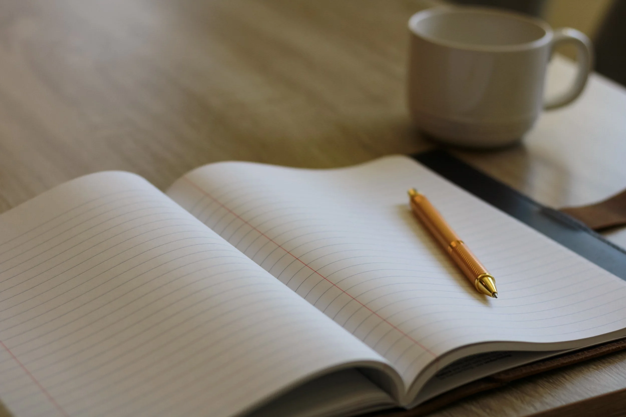 An open blank notebook with lined pages, a gold-colored pen resting on it, on a wooden table, with a white mug in the background.